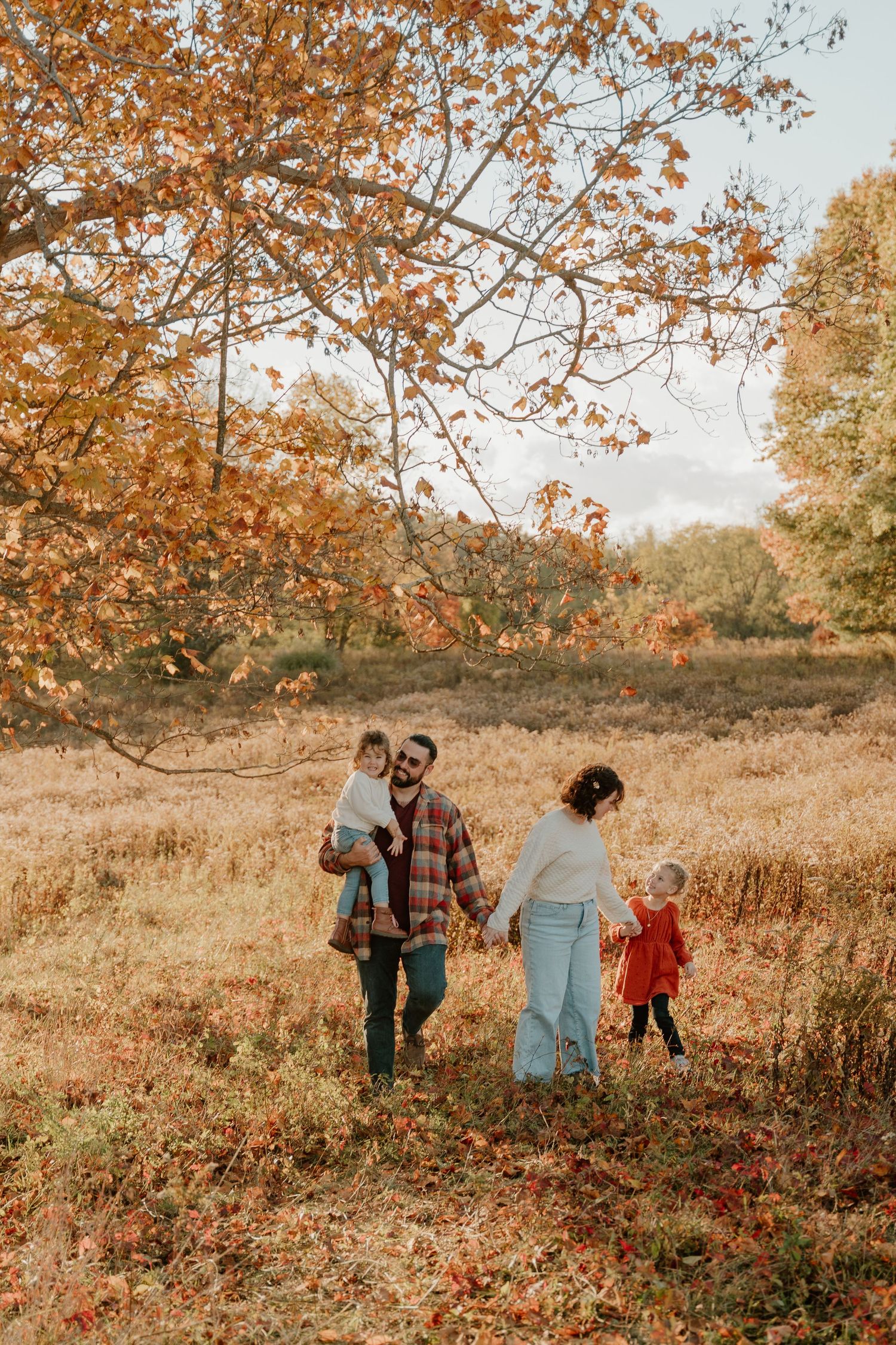 A family walks together through an autumn field with golden leaves and branches overhead on a warm fall day.
