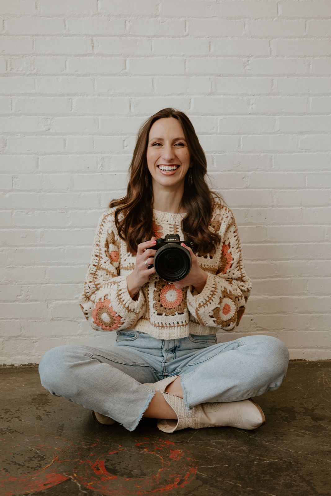 A person in a cozy sweater sits cross-legged against a white brick wall while holding a camera and laughing joyfully.