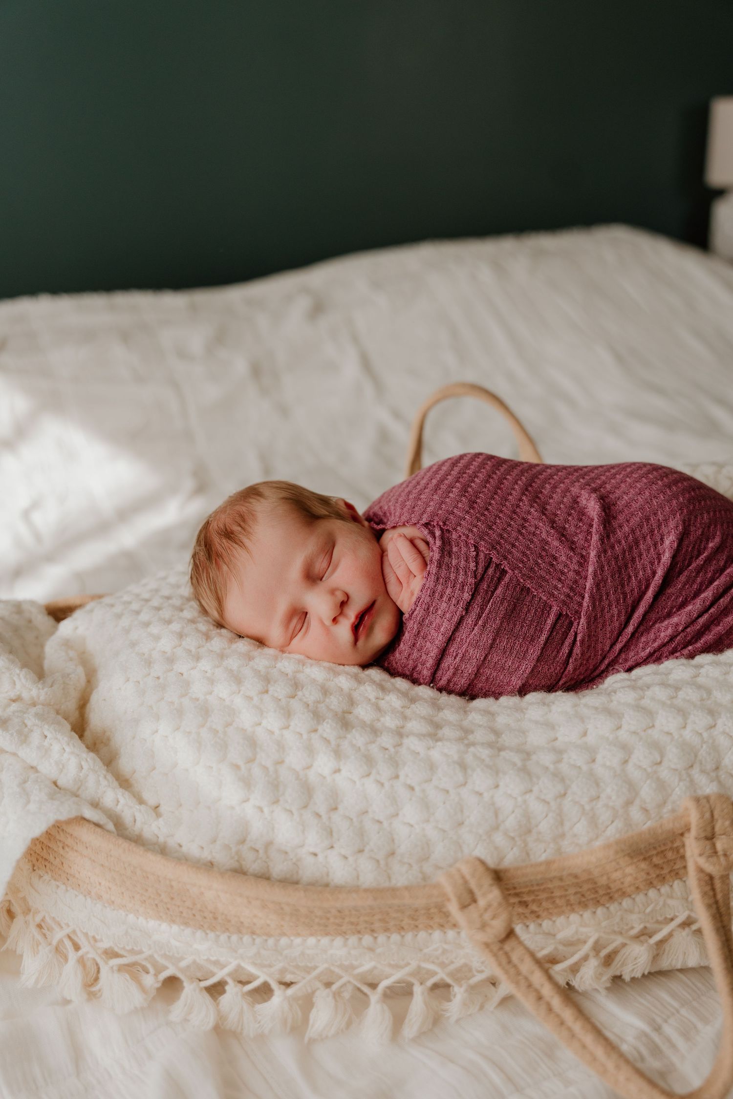 A sleeping baby wrapped in burgundy fabric lies peacefully on a textured white blanket.
