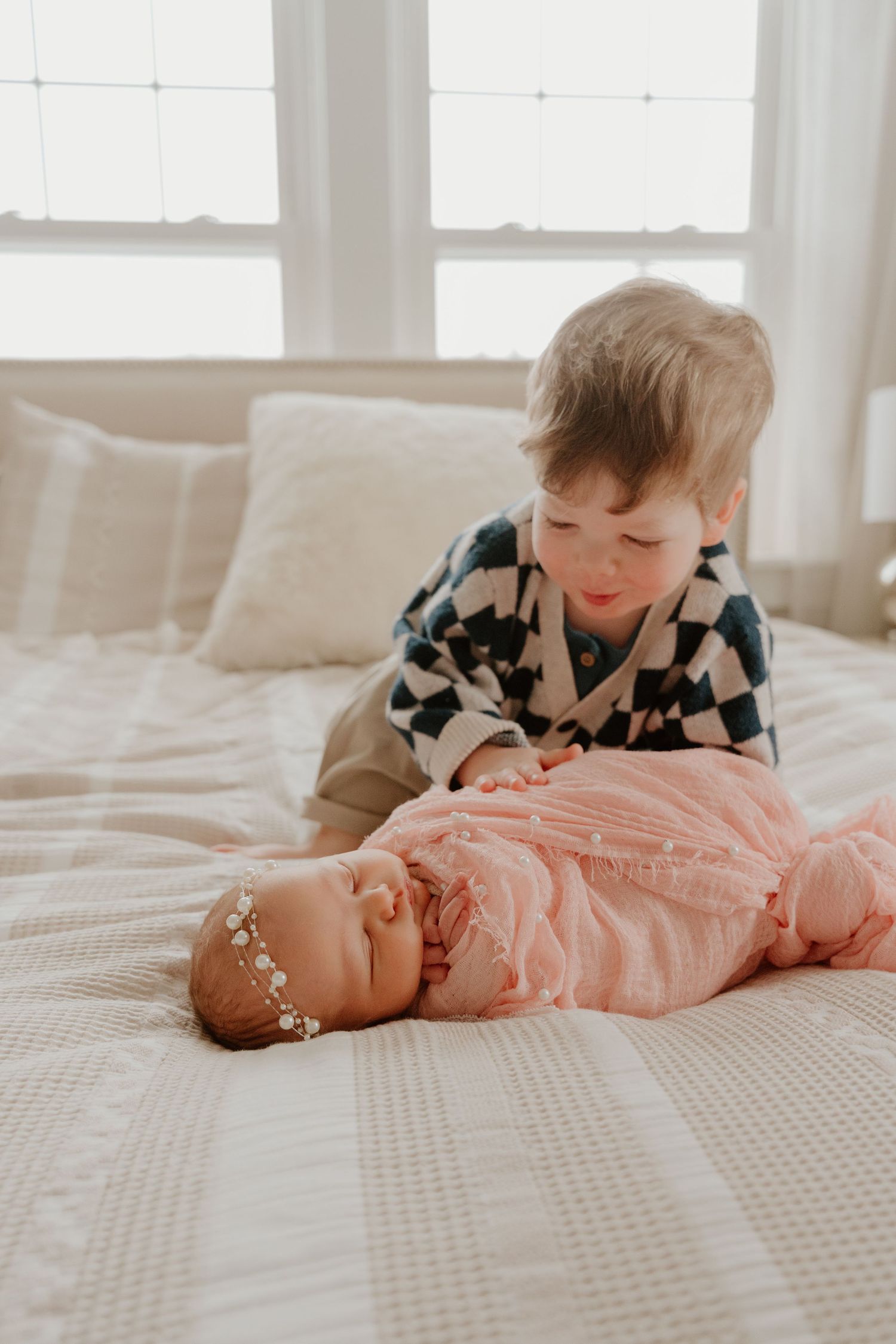 Two children interact sweetly with a baby wearing pink on a textured white bedspread.