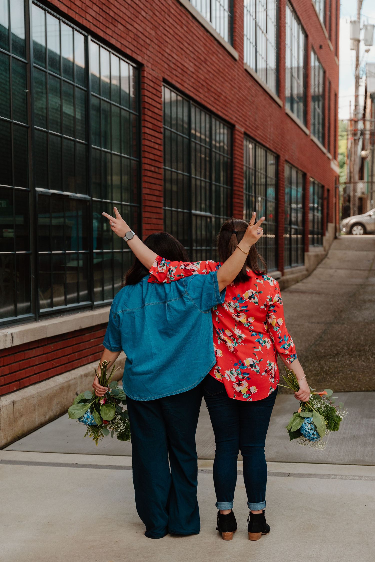 Two people in colorful shirts stand with their backs to camera holding flower bouquets against a brick building.
