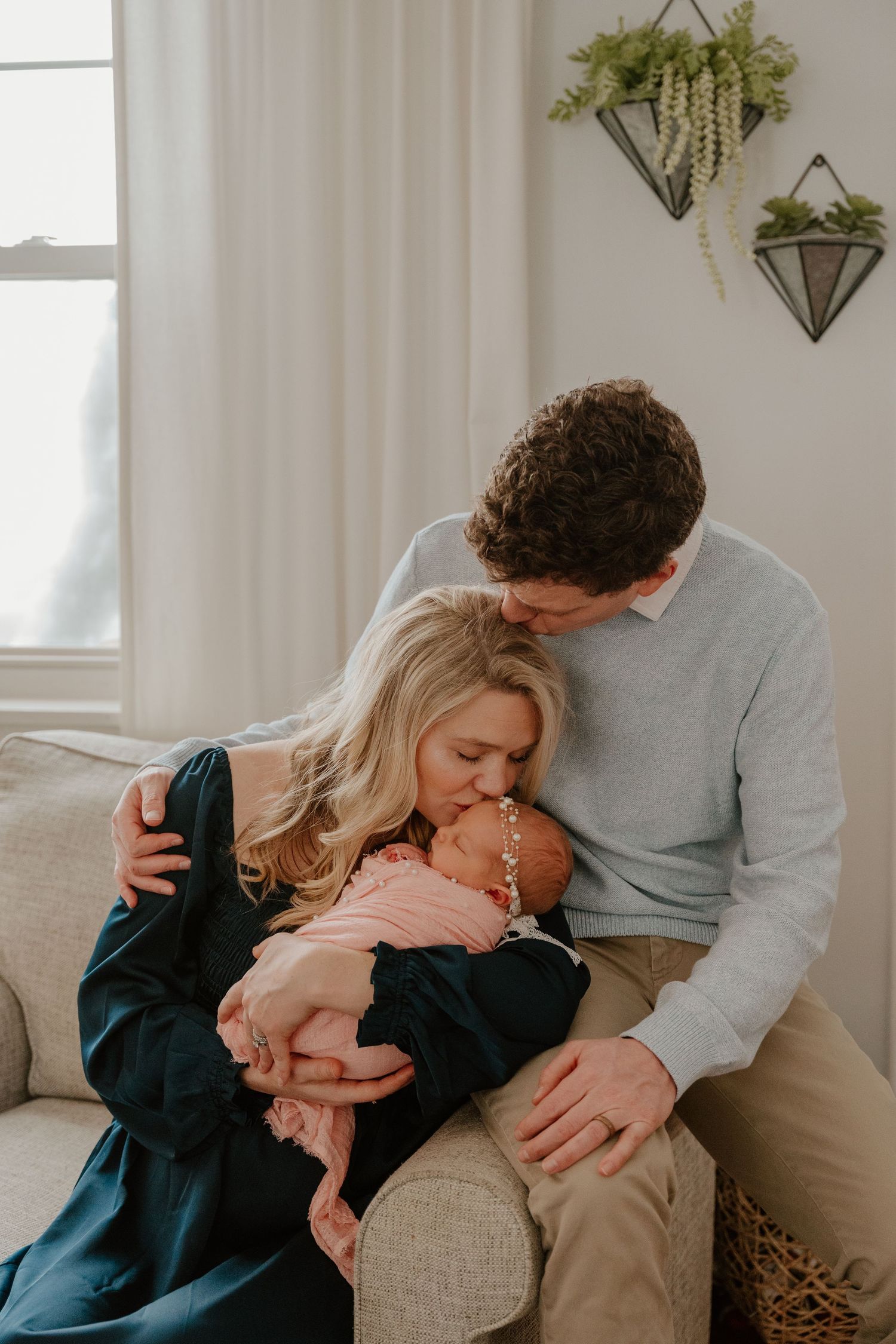 A family shares tender moments together on a couch in a bright living room with white curtains and wall decor.