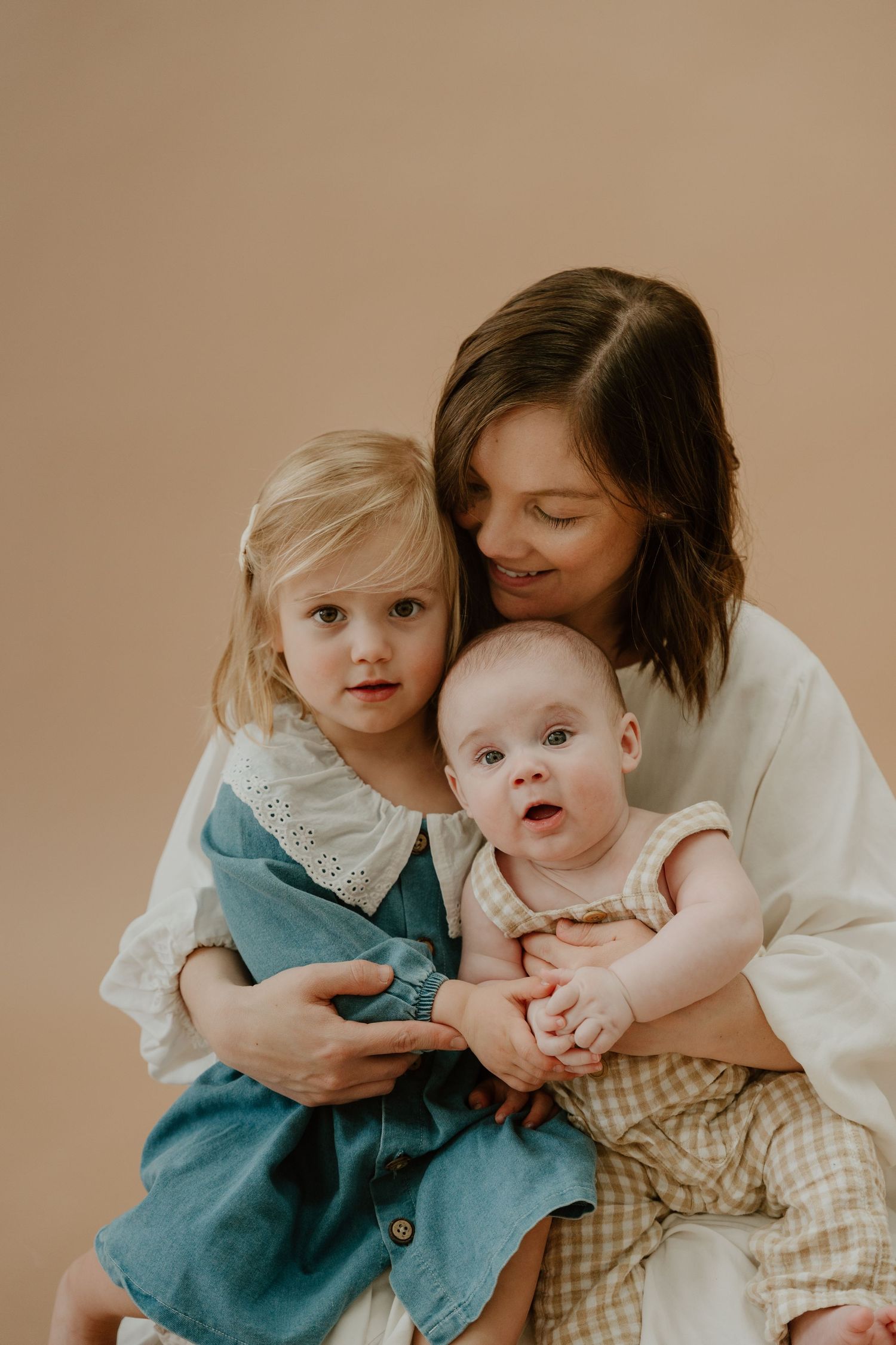 Intimate family portrait against beige backdrop featuring an infant and toddler in vintage-style clothing.