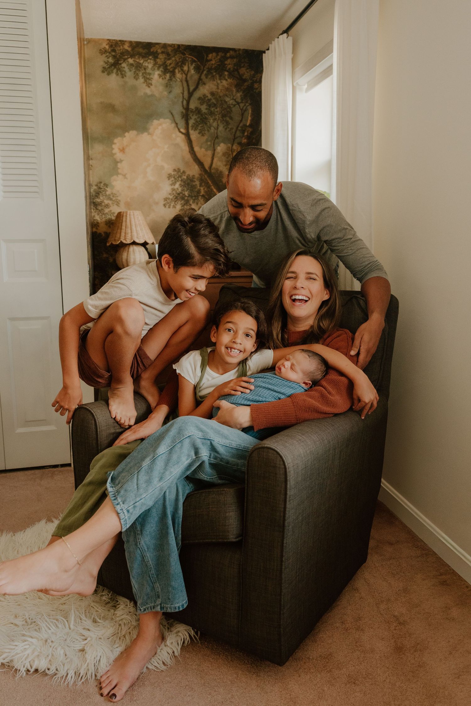 A family enjoys playful moments together on a grey armchair, sharing smiles and embraces in their living room.