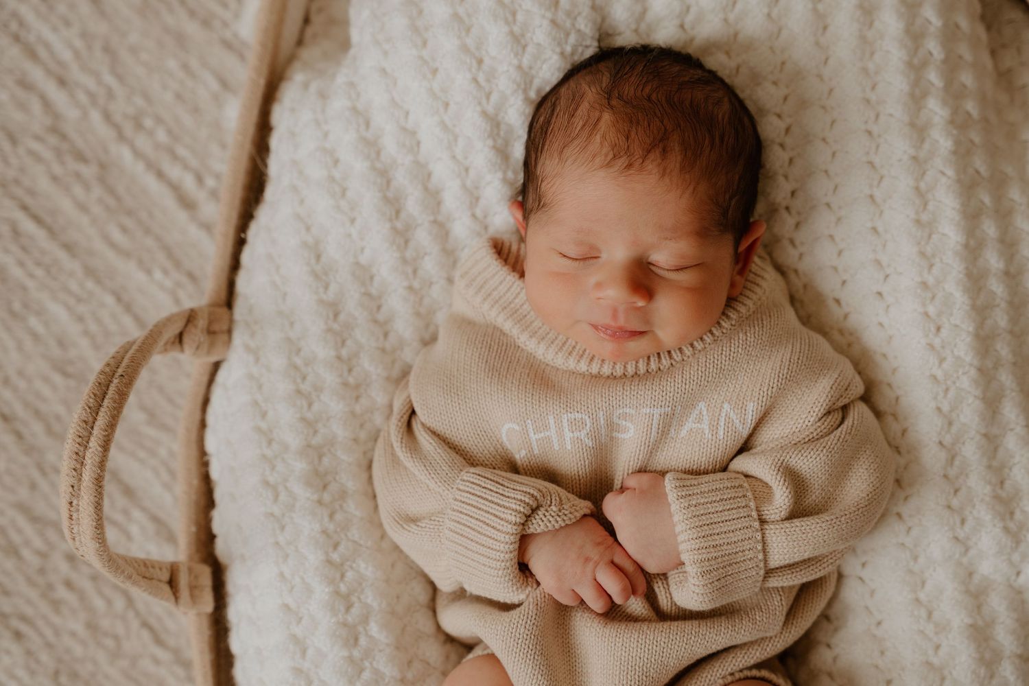 Sleeping newborn baby wearing cozy beige sweater lying on soft white blanket in natural light.