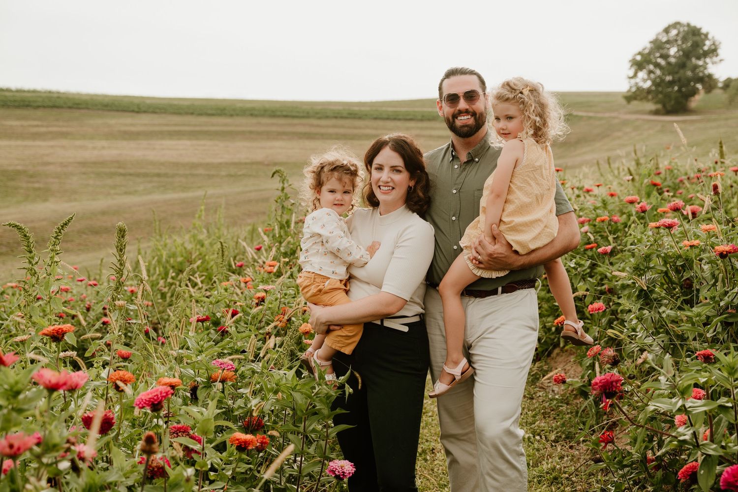 Family of four smiling in a flower field with red blooms and rolling green hills in the background.