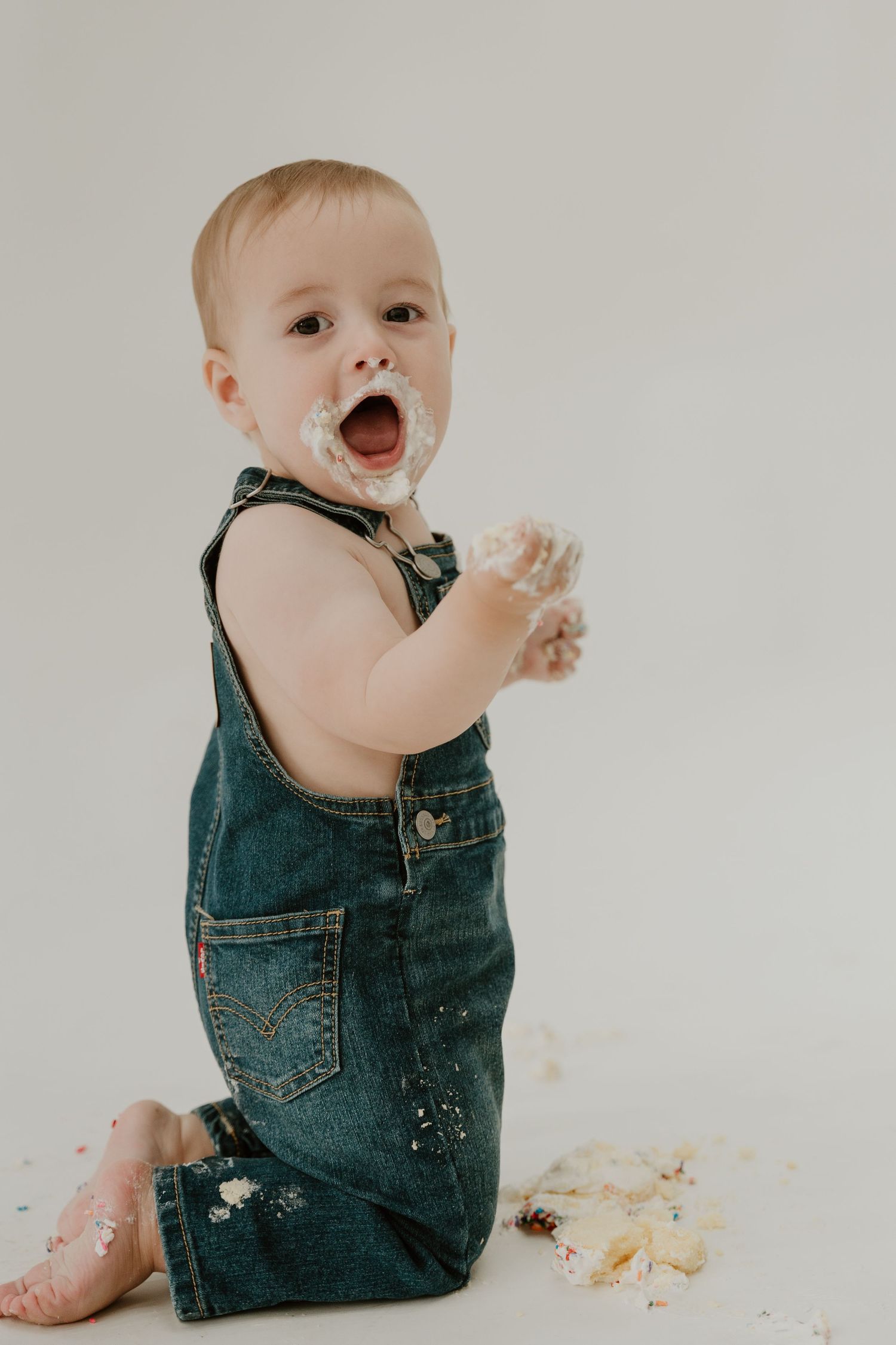 Baby in denim overalls sits on white background while messily eating cake and getting crumbs everywhere.