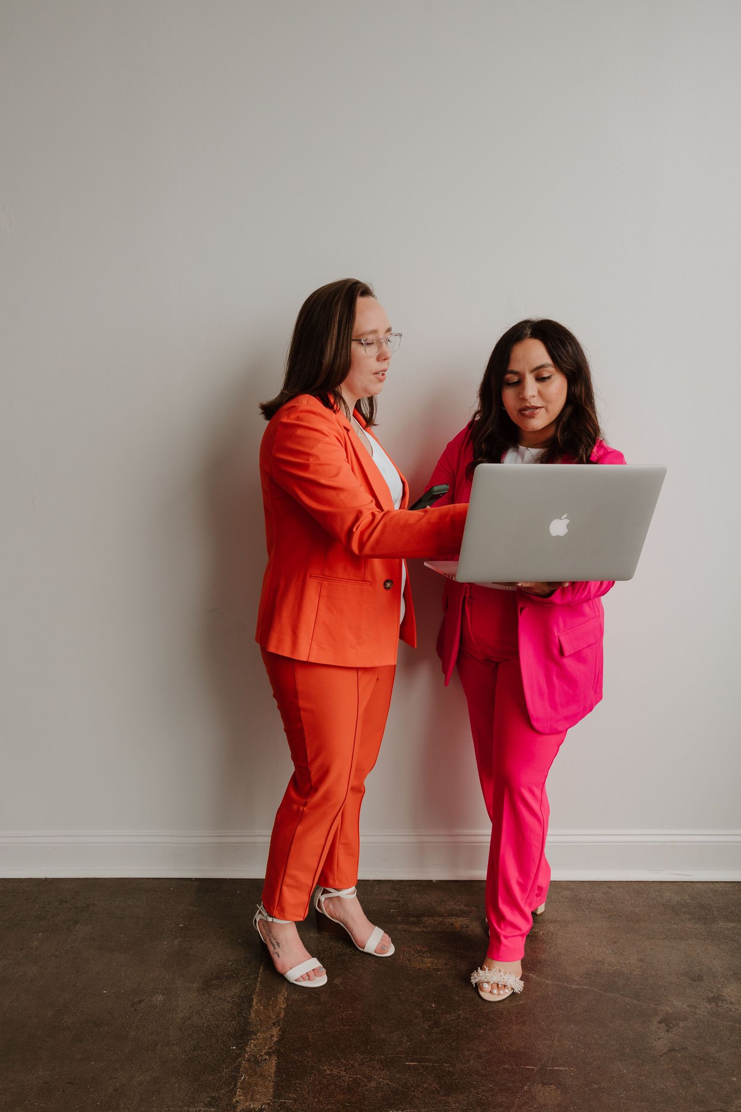 Two people in bright orange and pink outfits pose playfully with a laptop against a neutral background.