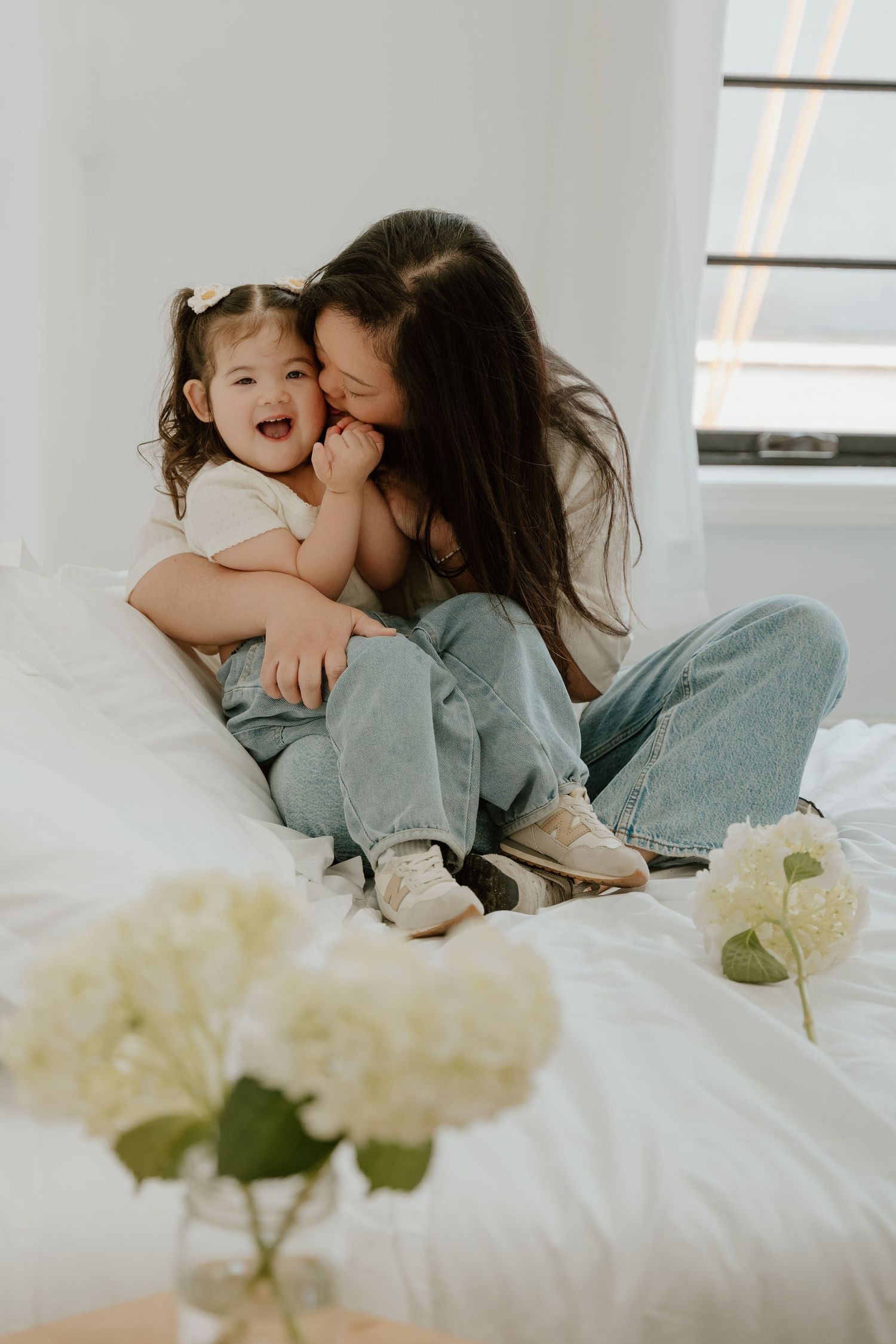 A joyful moment captured between parent and child laughing together on white bedding with hydrangeas in the foreground.