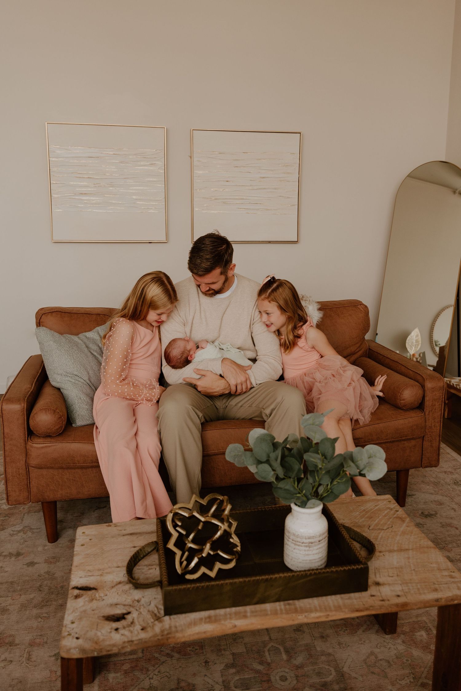 Family sits together on brown leather couch in cozy living room with decorative wall art and potted plant centerpiece.