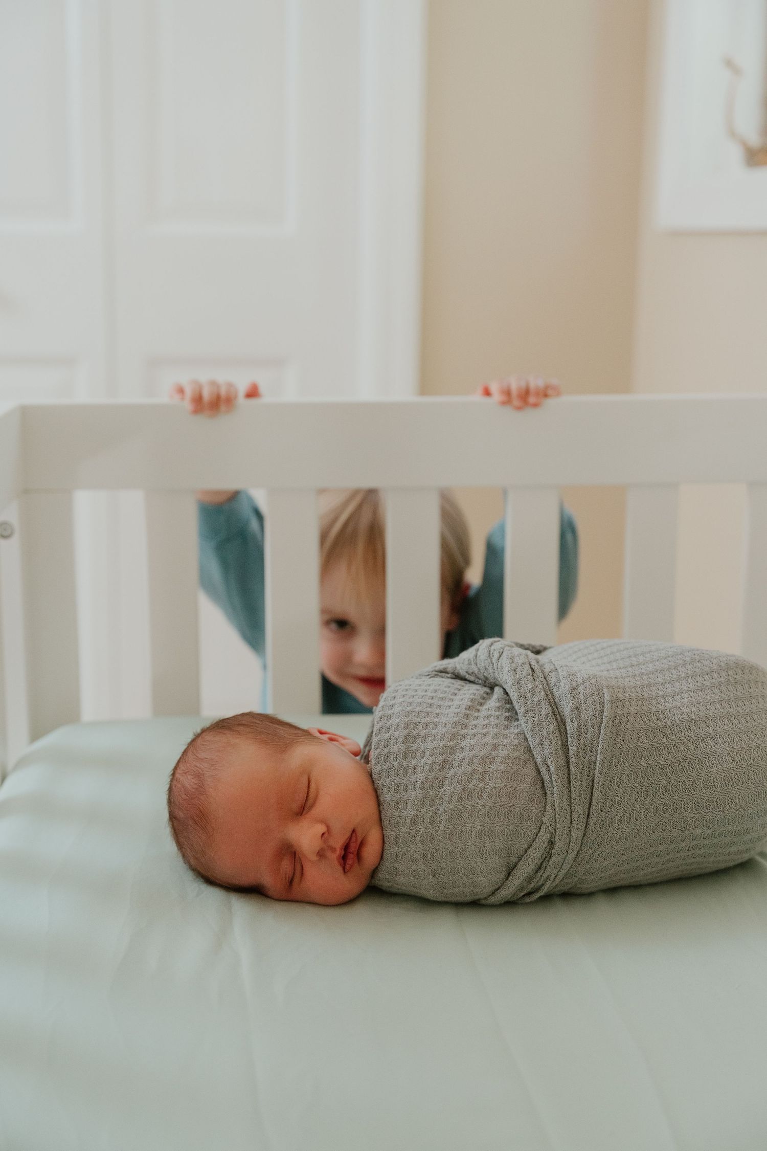 Sleeping baby wrapped in gray swaddle blanket lying in white crib viewed through slats.