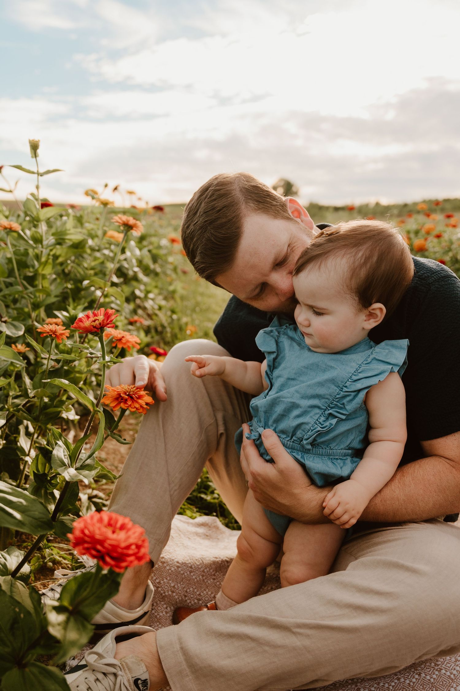 Father kissing baby girl while showing her red zinnia flowers in a vibrant outdoor flower field.