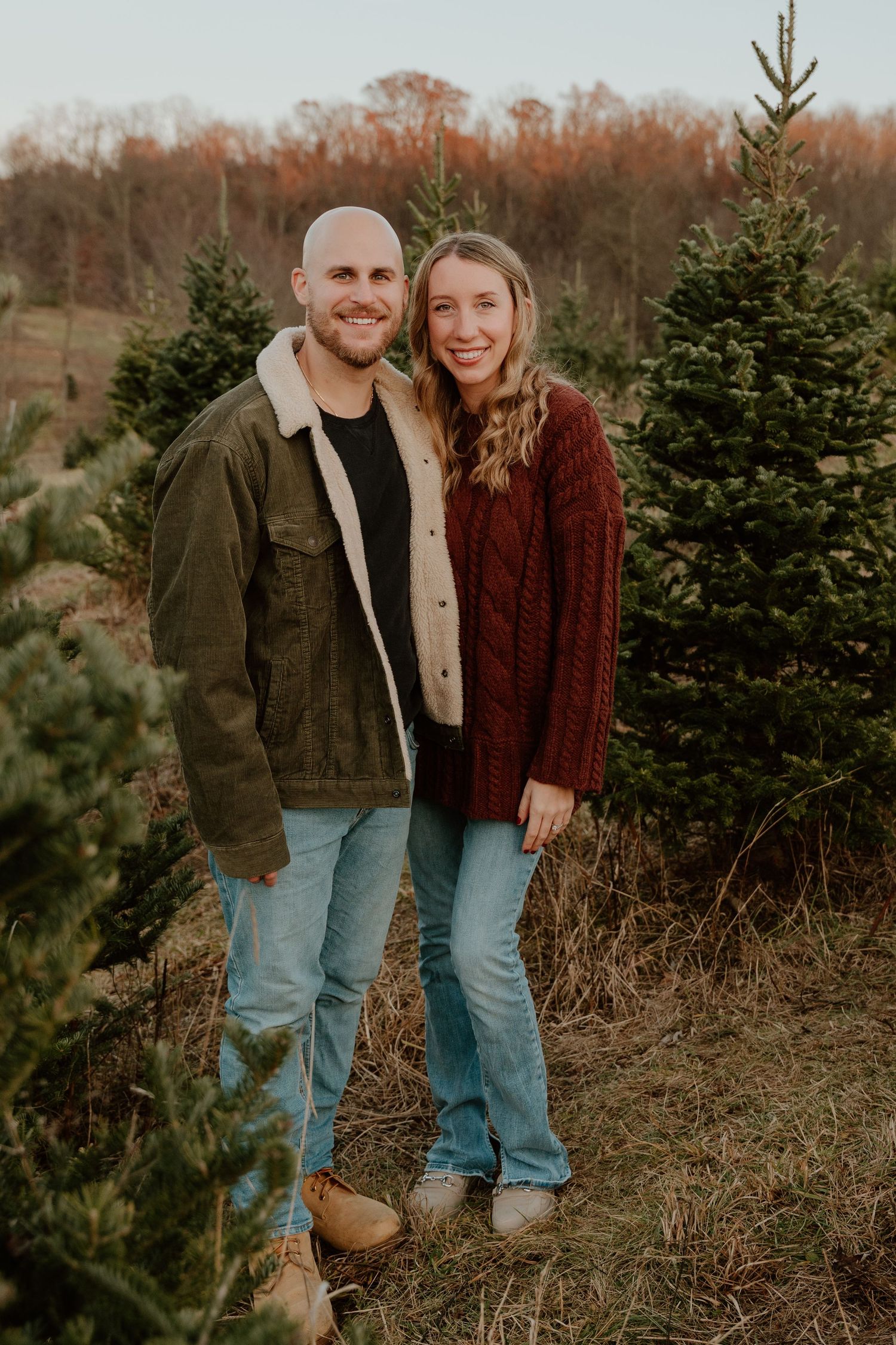 Couple in casual fall outfits pose together among evergreen trees during golden hour.