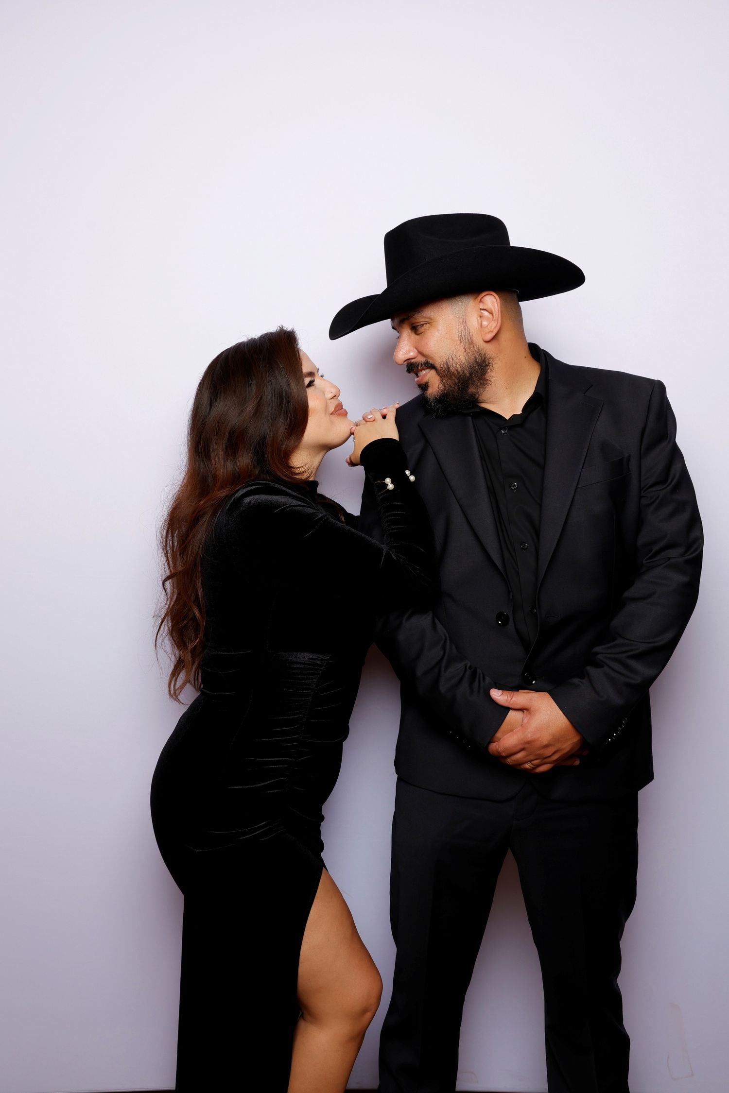Couple in all black formal wear share intimate moment against white backdrop.