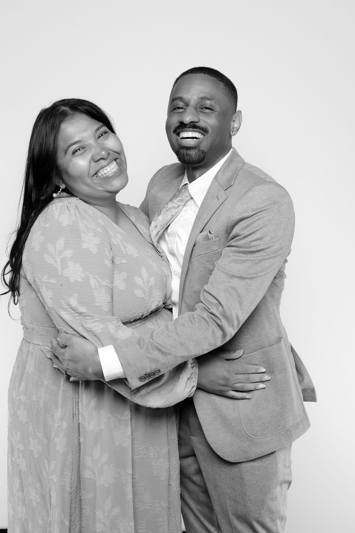 Black and white photo of a smiling couple embracing, man in suit and woman in floral dress.