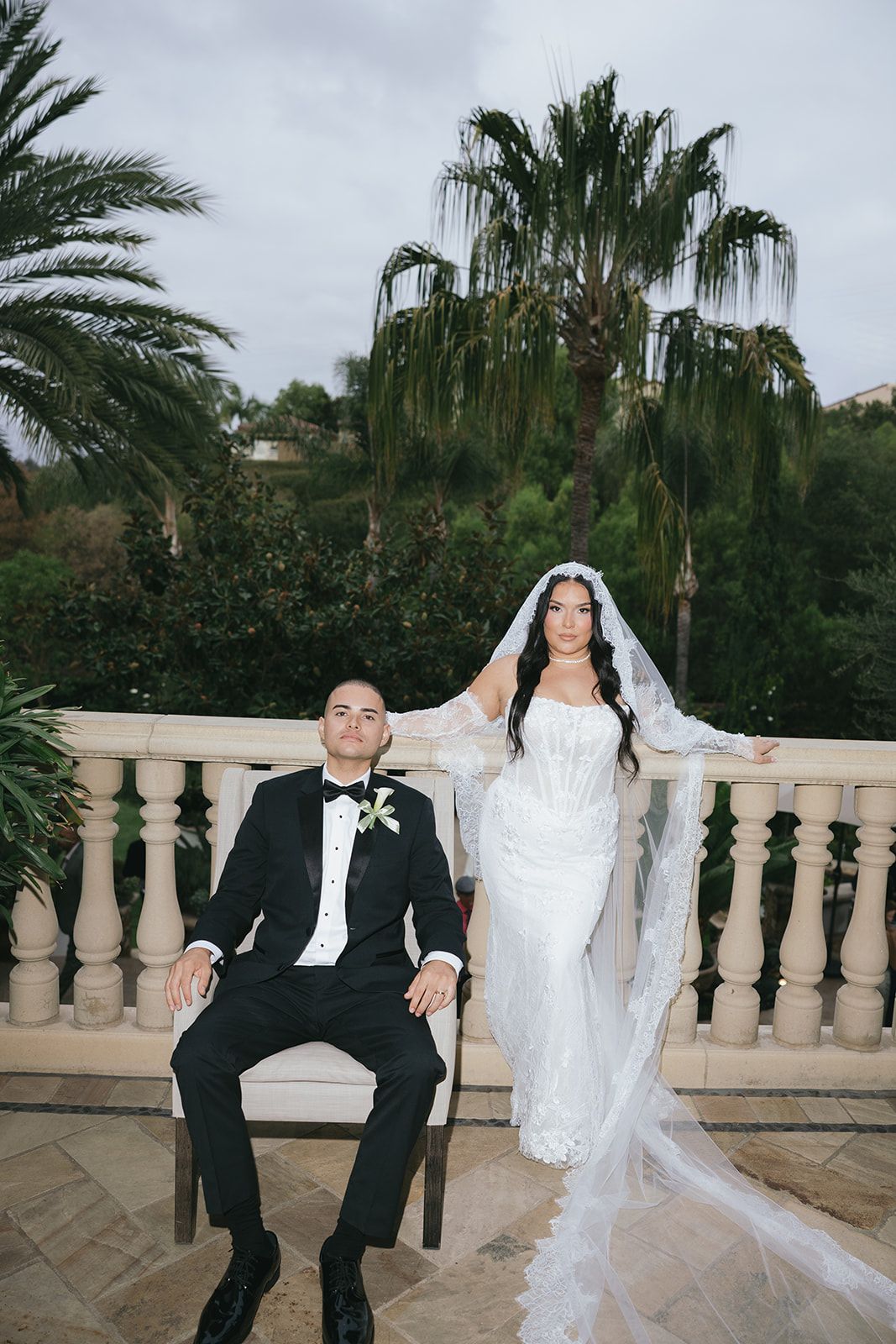 Wedding couple poses playfully on an elegant balcony with palm trees in the background.