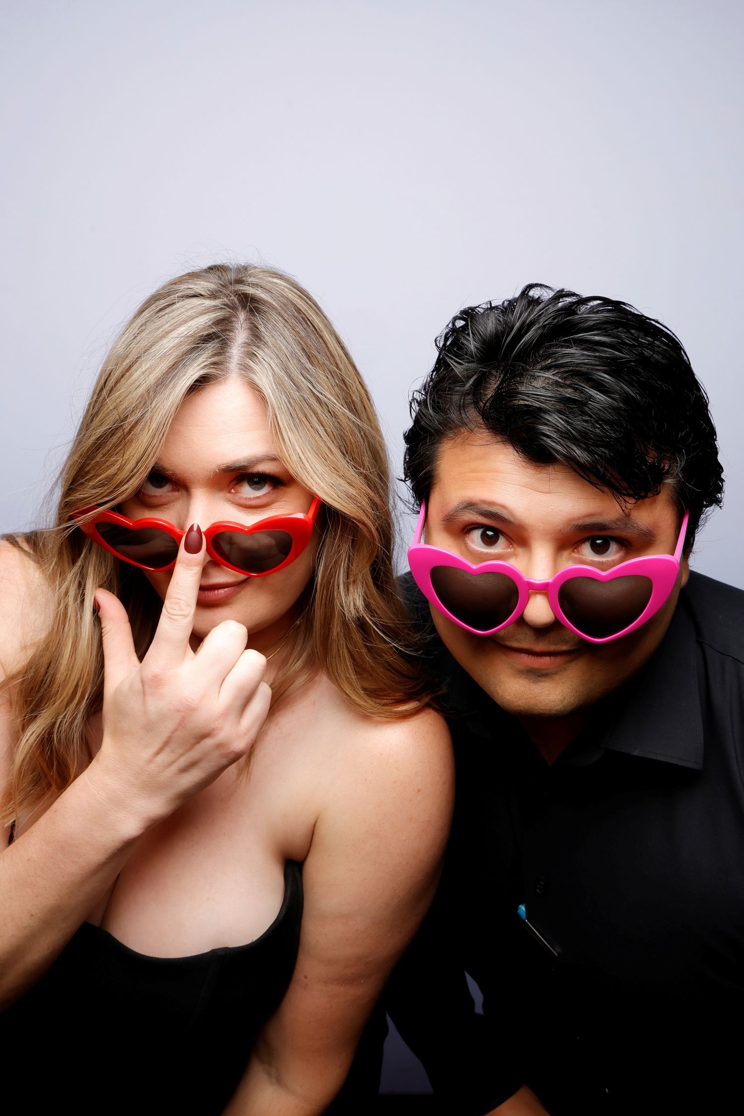 Two people wearing heart-shaped sunglasses pose playfully against a light studio background.