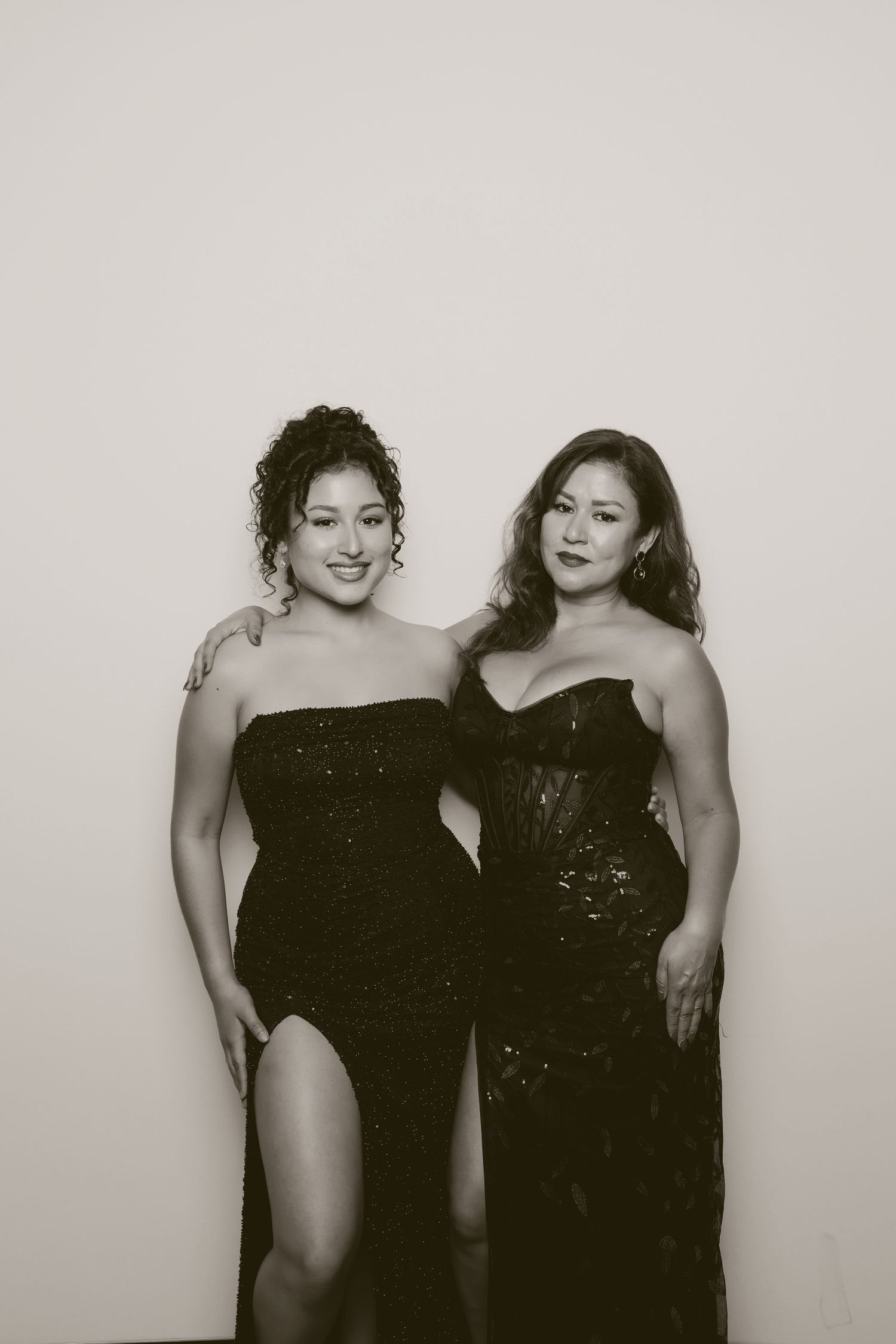 Two women in elegant black evening gowns pose together against light background.