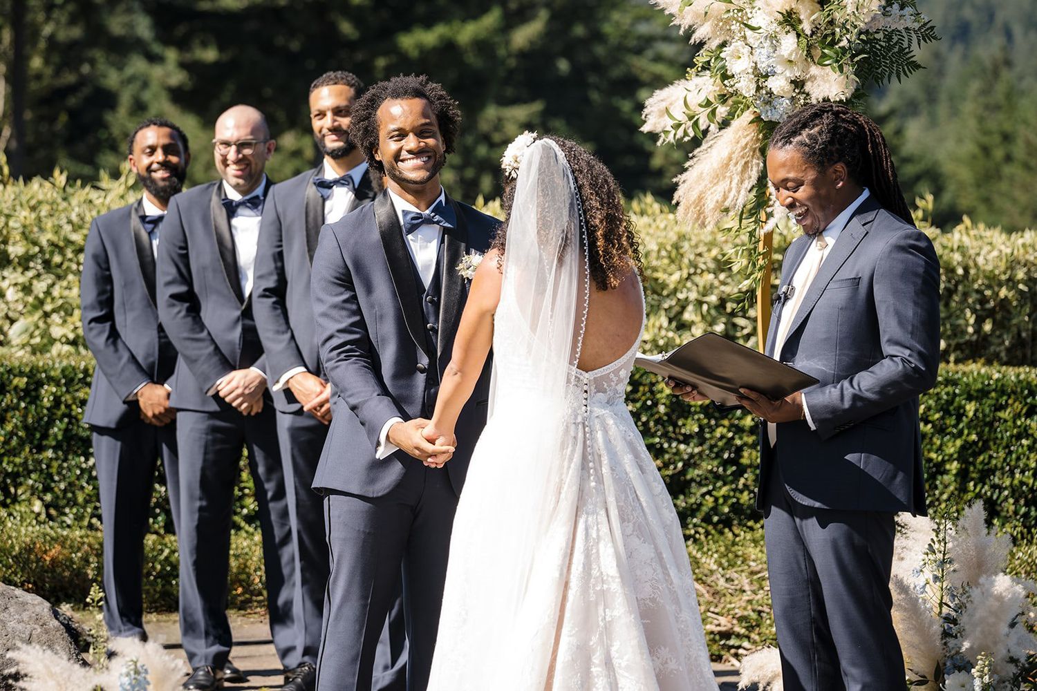 Outdoor wedding ceremony with groom in blue suit holding brides hands before smiling officiant and groomsmen.