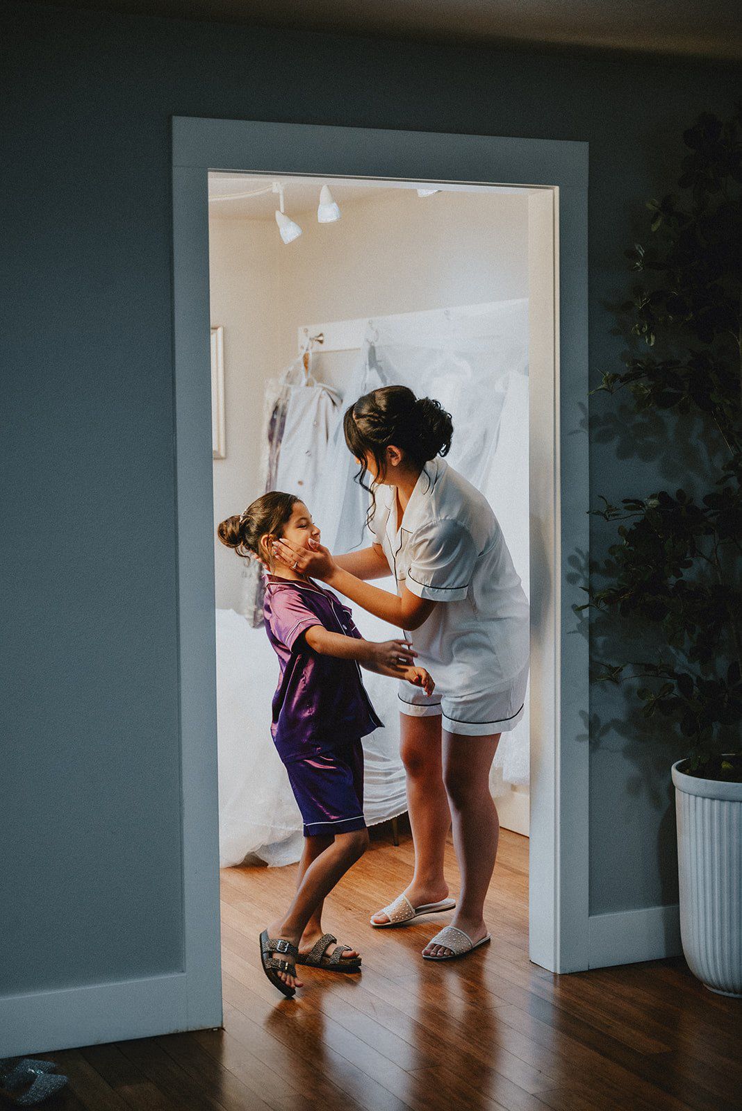 Woman helping a young girl get ready in a bright bedroom doorway, with a wedding dress hanging in the background.