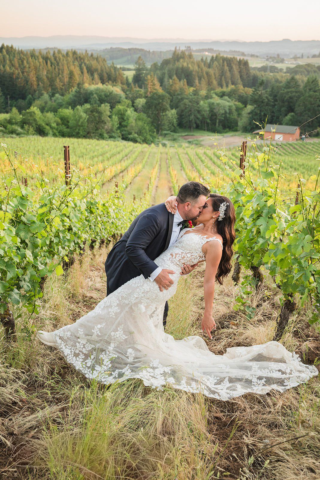 Bride and groom share a romantic dip kiss among lush vineyard rows with rolling hills at sunset.