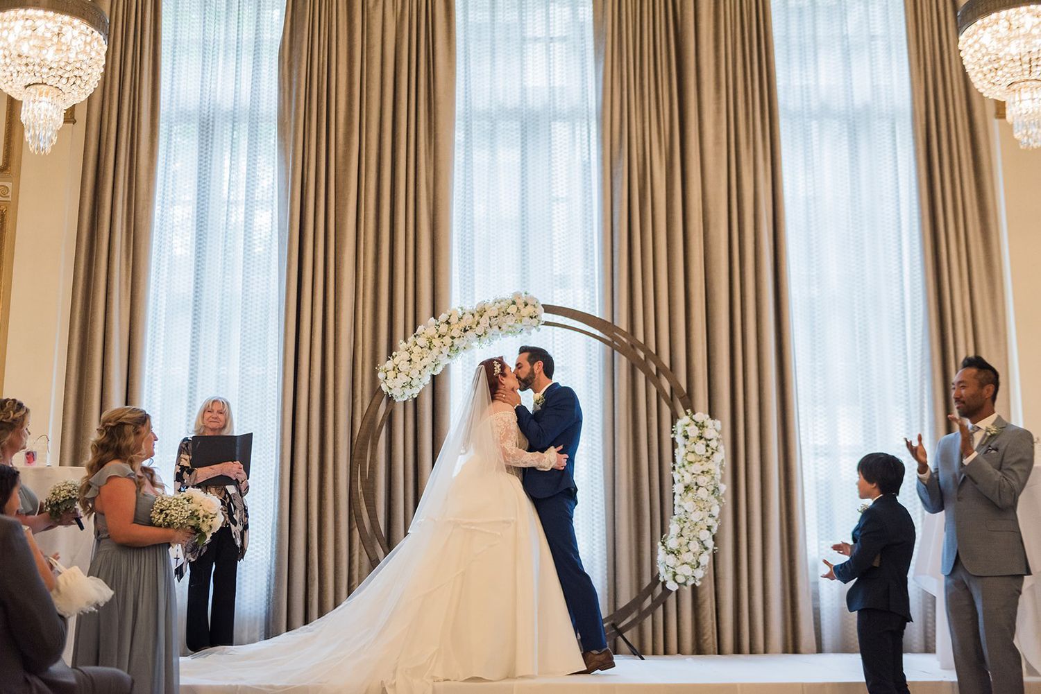 Bride and groom share first kiss under floral arch at elegant wedding ceremony with tall draped windows.