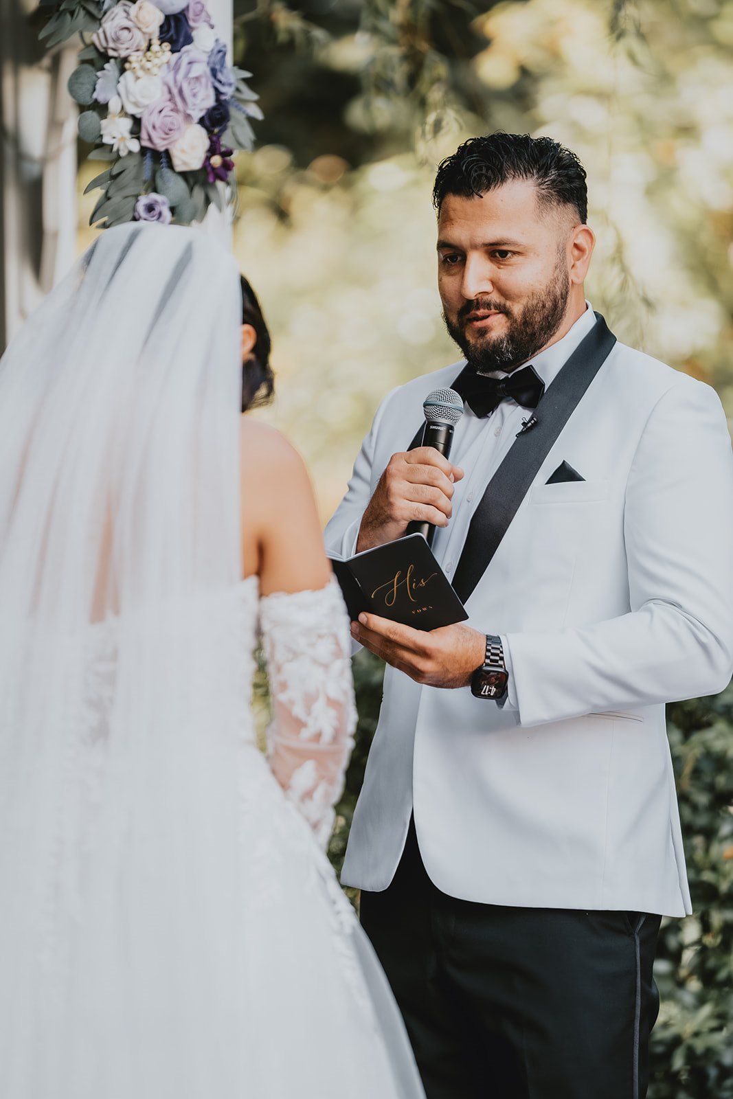 Groom in white tuxedo reads vows from black book at outdoor wedding ceremony while holding microphone.