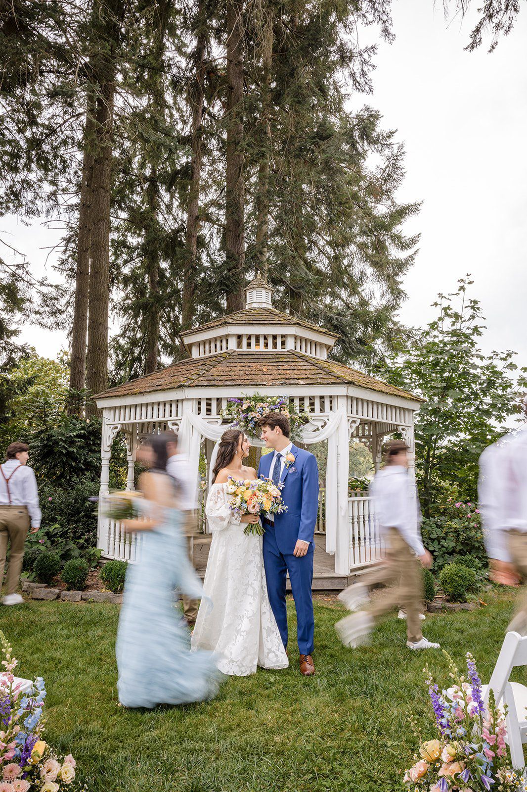 Bride and groom stand before a white gazebo outdoors, surrounded by blurred guests and tall trees at a garden wedding.