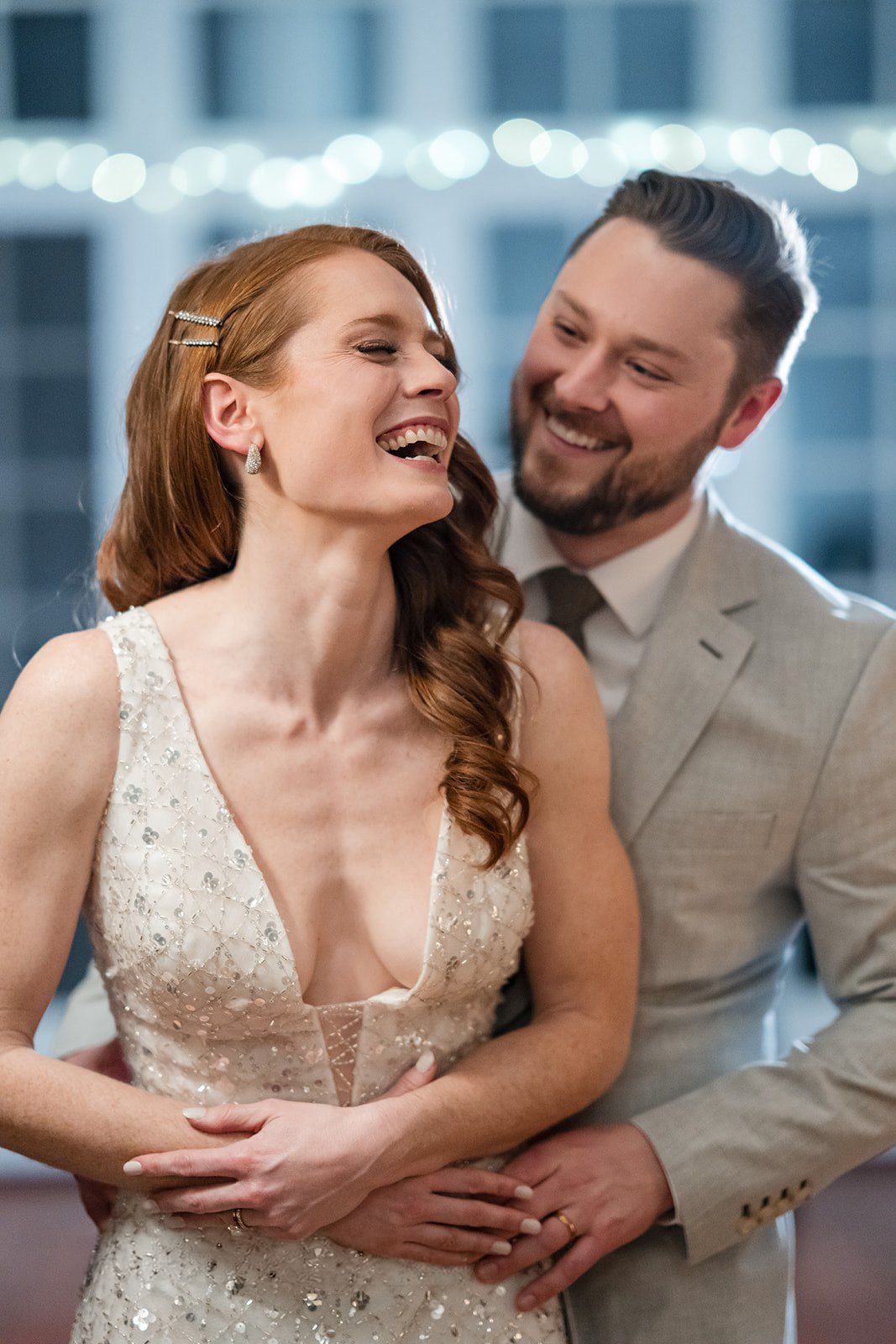 Laughing redhead bride in beaded gown embraced by smiling groom in tan suit at evening reception.
