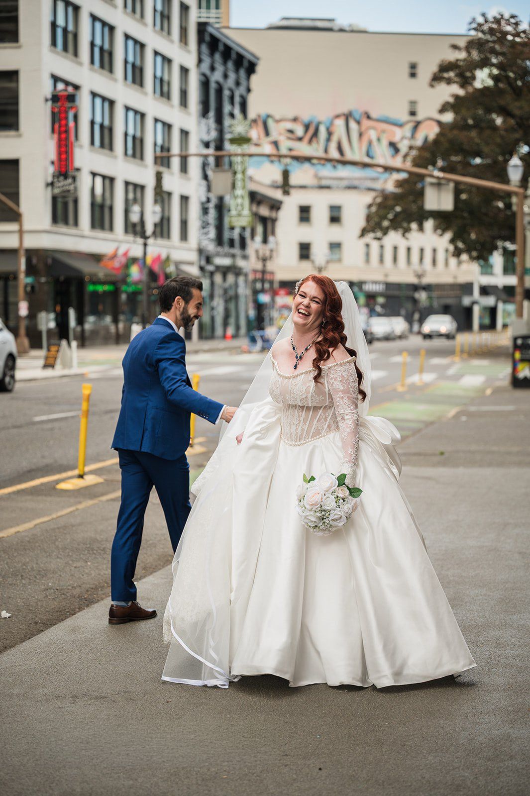 Bride in white ball gown and groom in blue suit hold hands while crossing a city street, with urban buildings behind them.