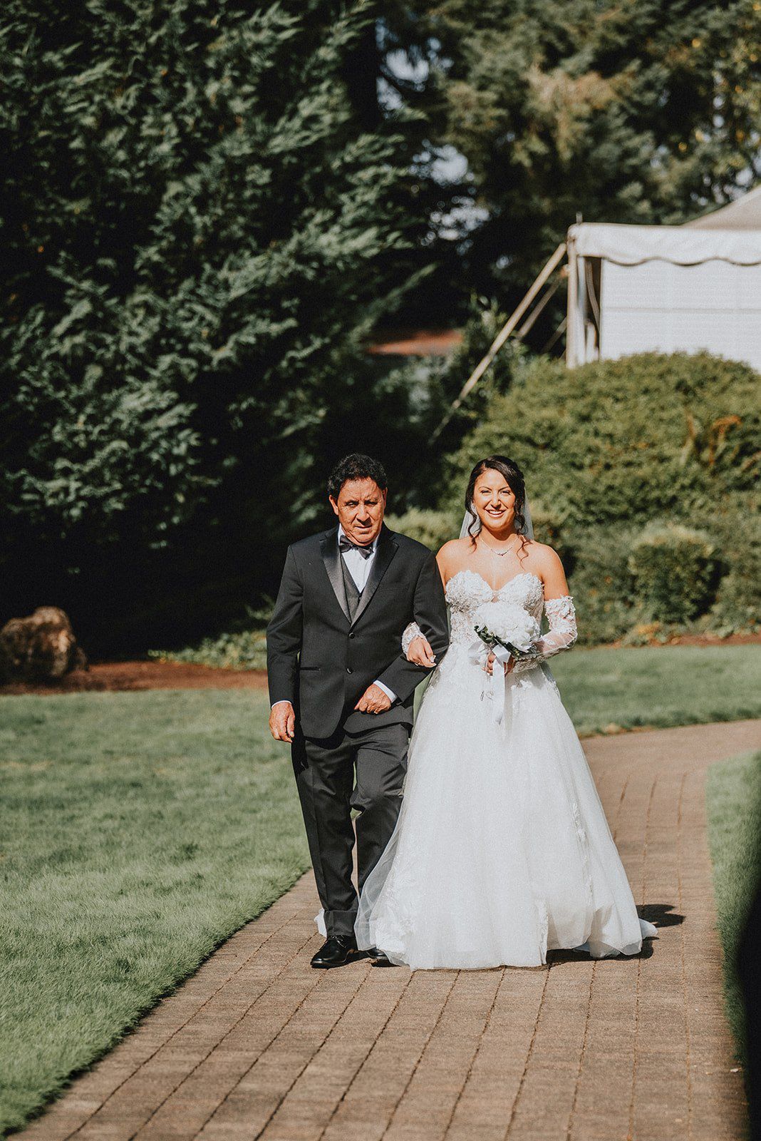 Bride in white ball gown walks arm-in-arm with suited man on brick path through lush garden on wedding day.