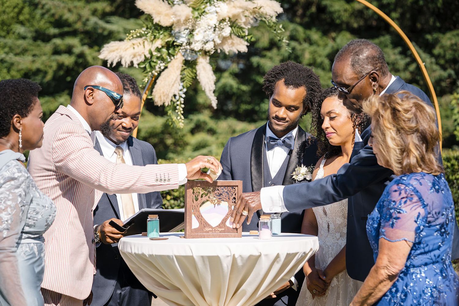 Wedding guests gather around a decorated table during an outdoor ceremony under a floral arch.