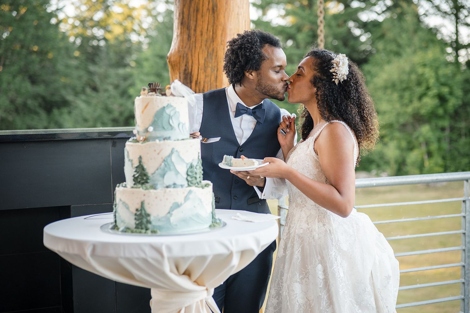 Newlywed couple sharing a kiss while cutting a elegant three-tier mountain-themed wedding cake outdoors.