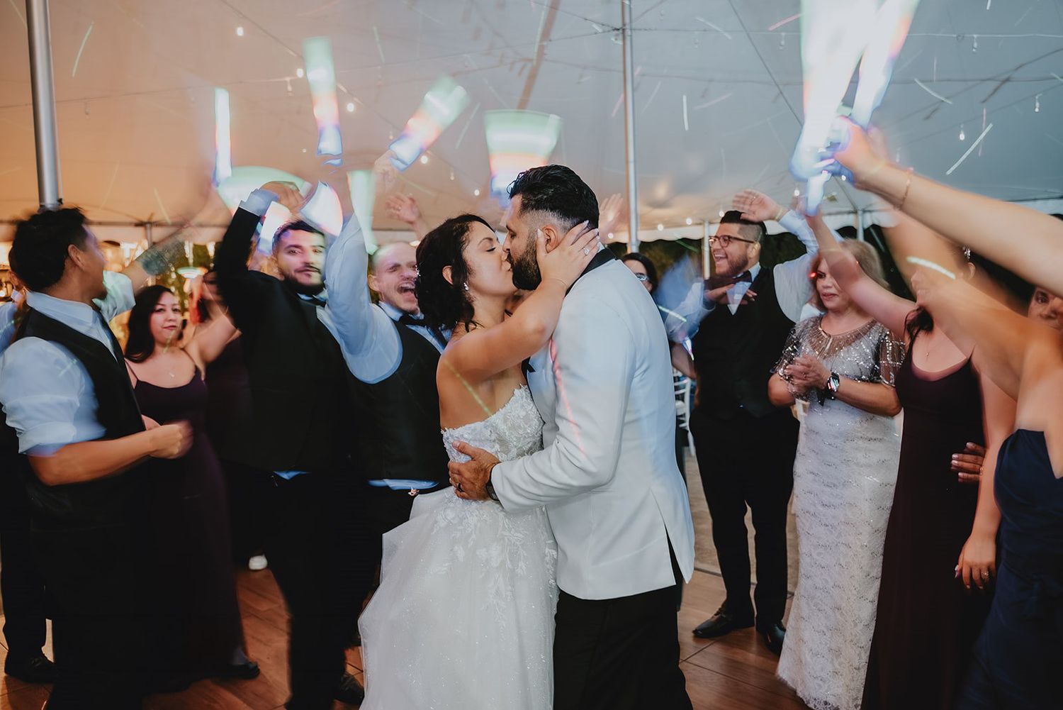 Newlywed couple shares first dance kiss surrounded by cheering guests waving sparklers at elegant wedding reception.