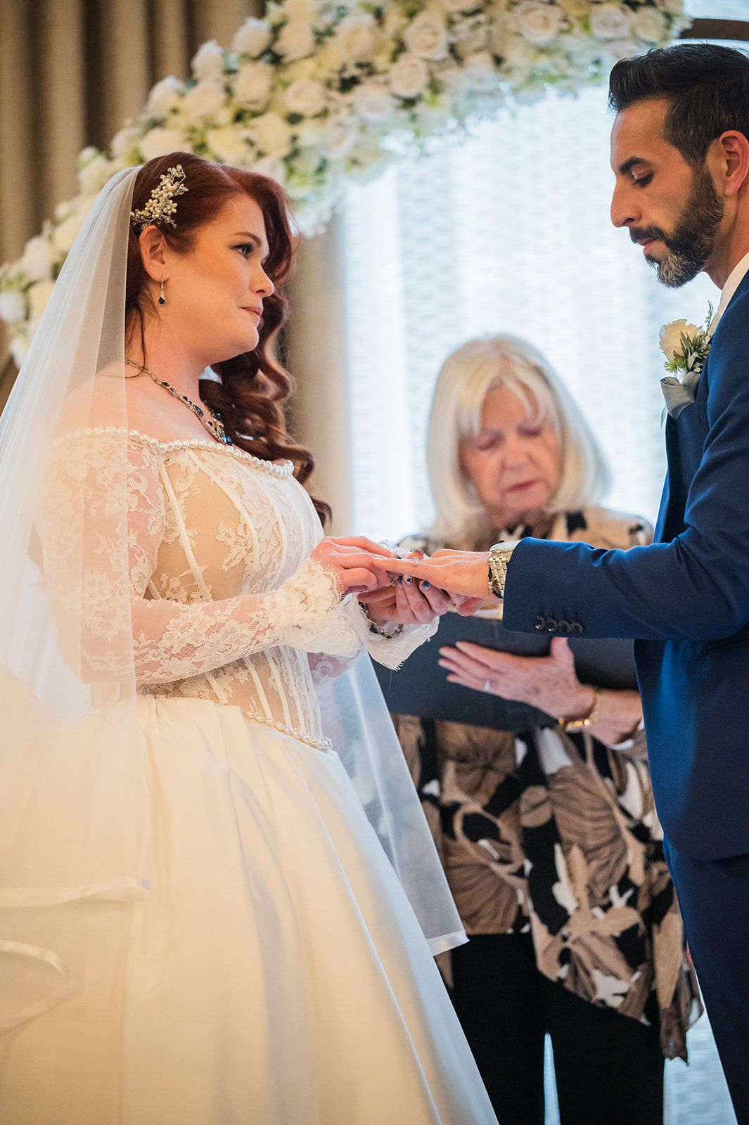 Bride placing wedding ring on groom's finger during ceremony, officiant in background, floral arch.
