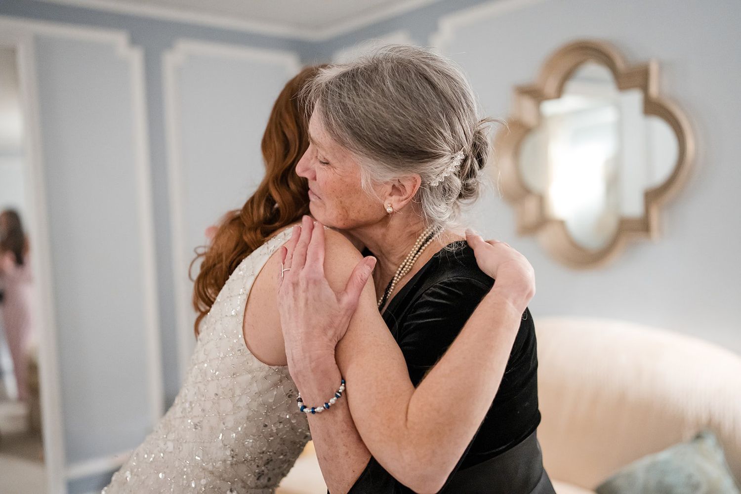 A bride in a beaded gown shares a heartfelt hug with an older woman in a black dress during wedding preparations.