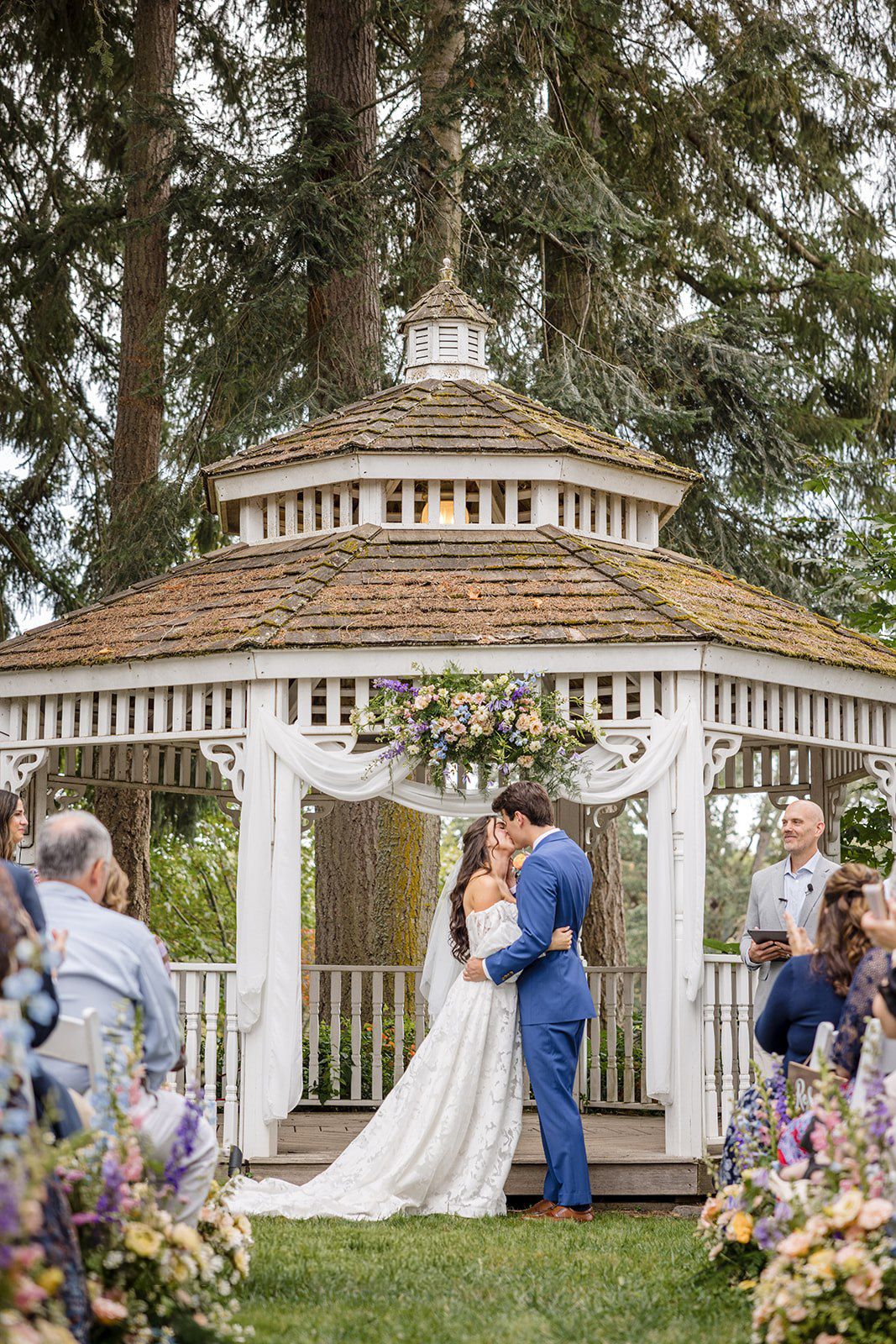 Couple shares first kiss under a white gazebo adorned with colorful flowers during an outdoor wedding ceremony.
