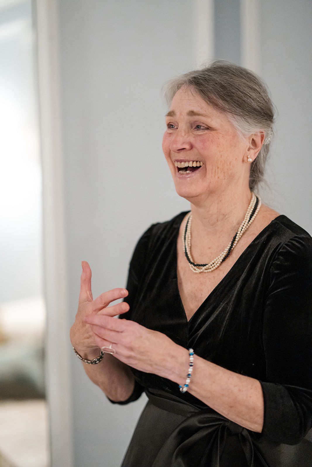 Elderly woman in black velvet dress laughing joyfully, wearing pearl necklace and bracelet.