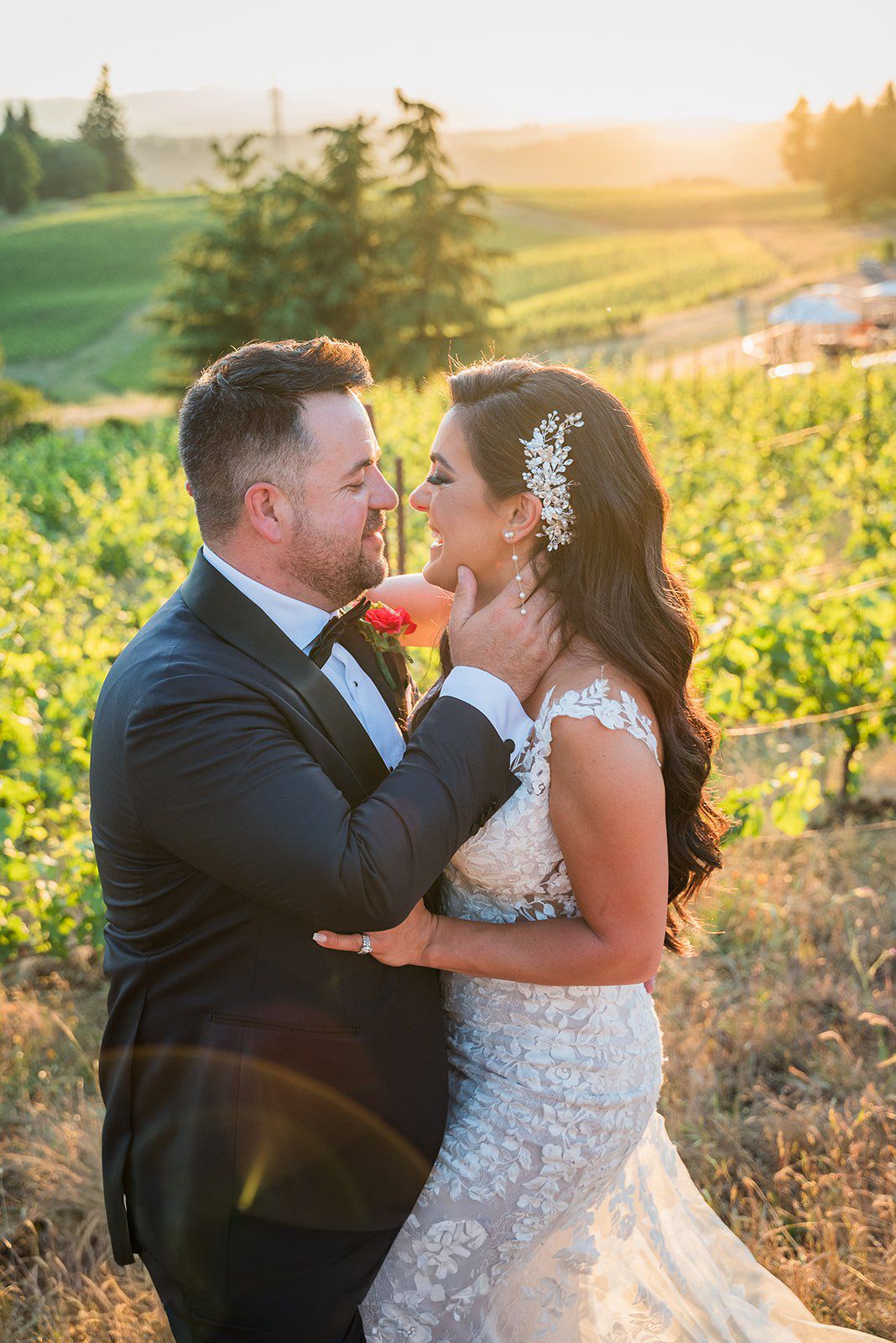 Bride and groom share a romantic moment at sunset in a lush vineyard, bride wearing lace gown with floral hair piece.