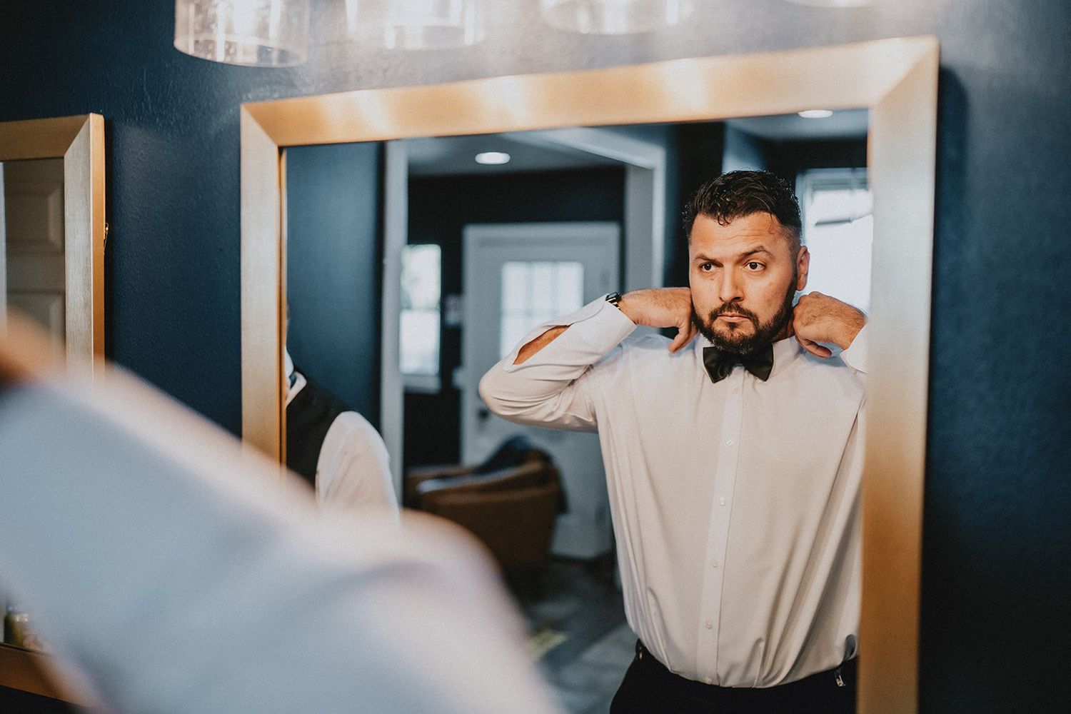 Groom adjusting his black bow tie while looking in a mirror during wedding preparations.