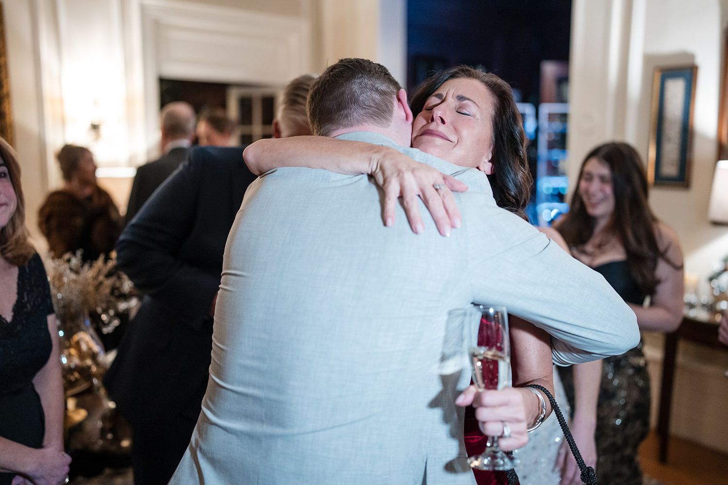 Two guests share a warm hug at an elegant indoor party, woman holding a wine glass, others socializing in background.