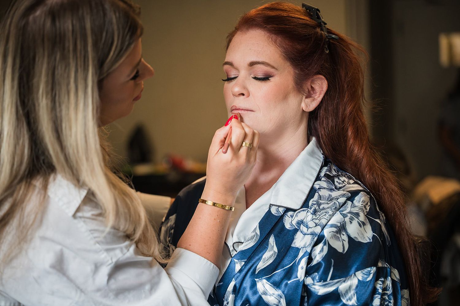 Makeup artist applying red lipstick to a woman with long auburn hair in a floral robe.
