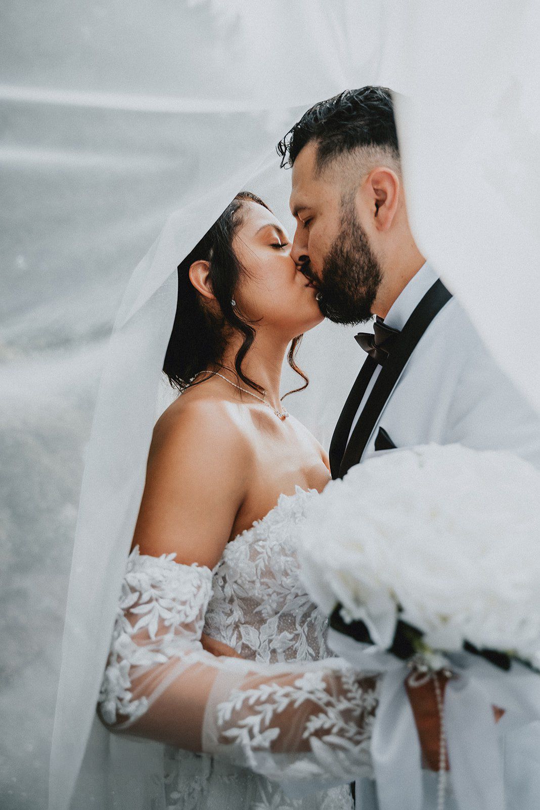 Bride and groom sharing a romantic kiss under a flowing white veil, bride holding a white bouquet in lace off-shoulder gown.