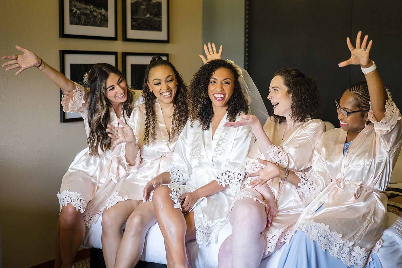 Five bridesmaids in matching blush satin robes celebrate joyfully with the bride wearing a veil on a wedding morning.
