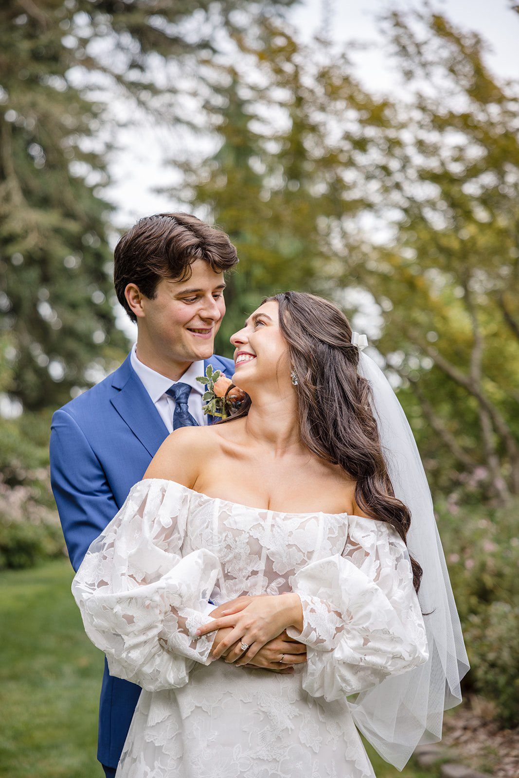 Smiling bride in off-shoulder lace gown and groom in blue suit share a romantic moment outdoors.
