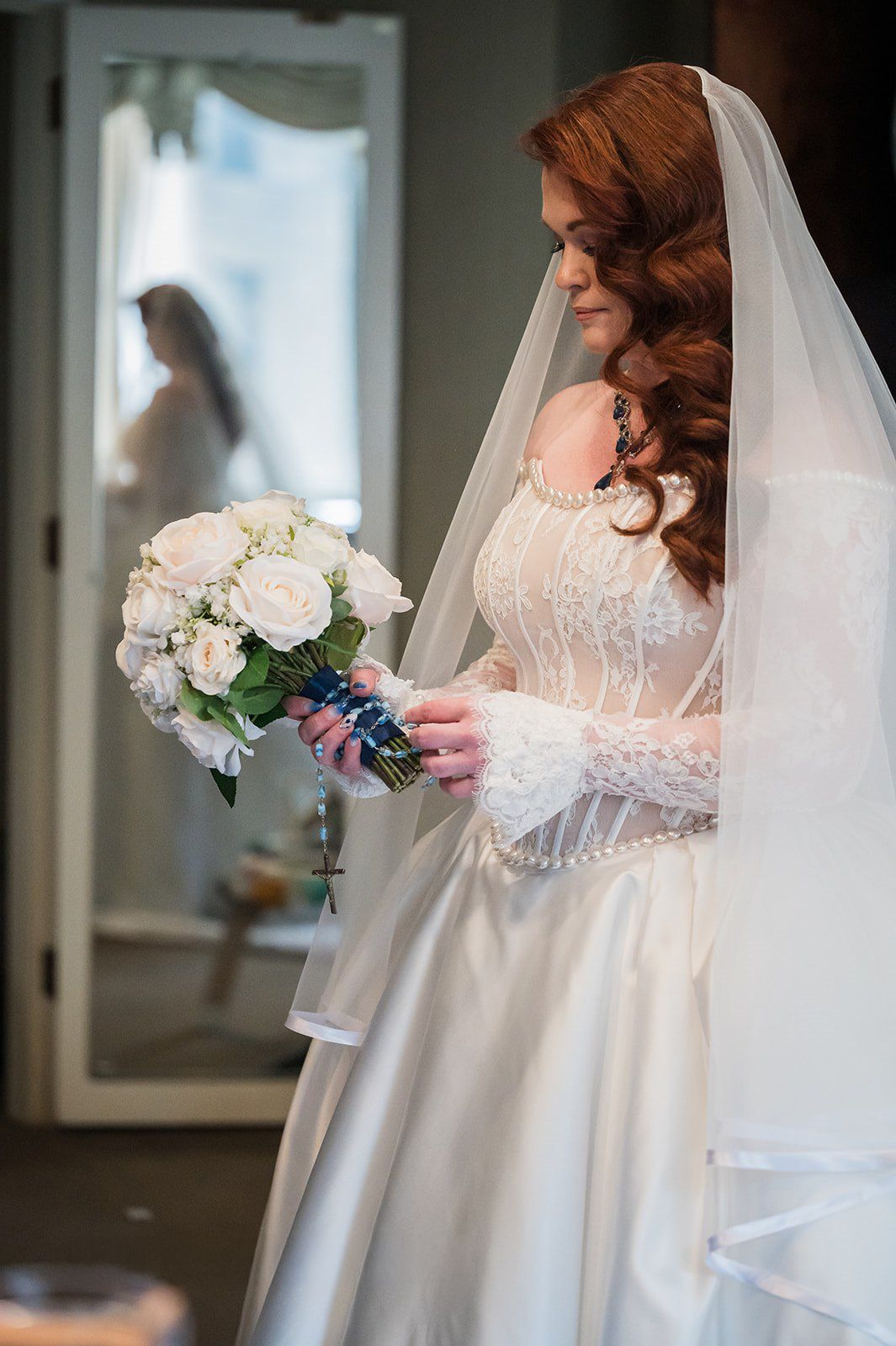 Red-haired bride in white lace gown and veil holds white rose bouquet, with mirror reflection in background.