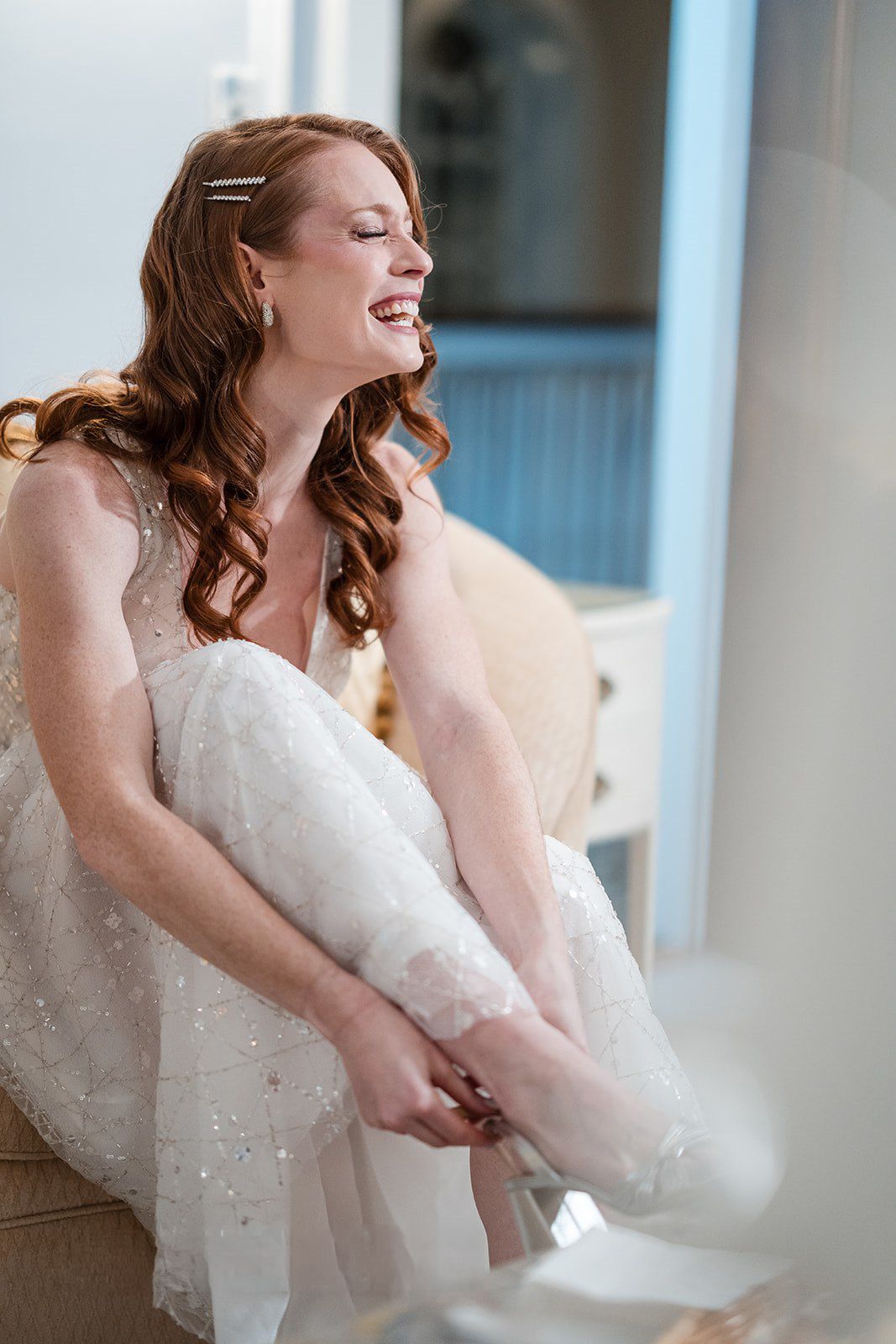 Smiling bride in a sparkly white gown adjusting her shoe while seated, with curly red hair and hair pins.