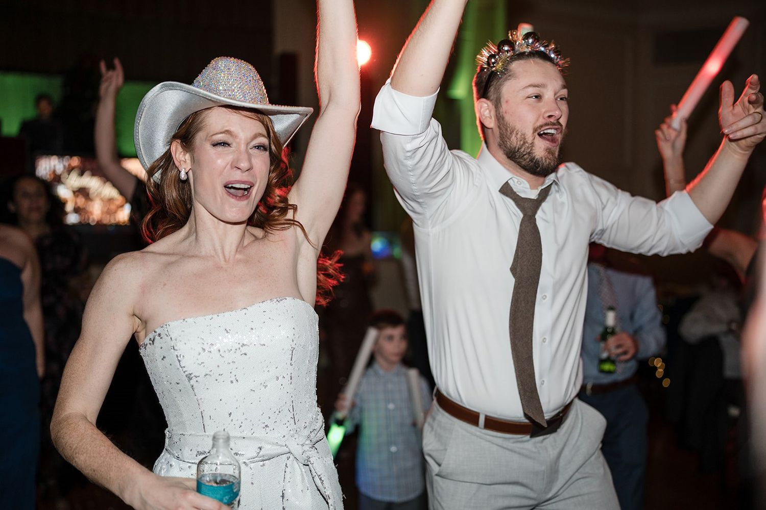 Bride and groom dancing joyfully at wedding reception, bride in white dress with cowboy hat, groom in white shirt.