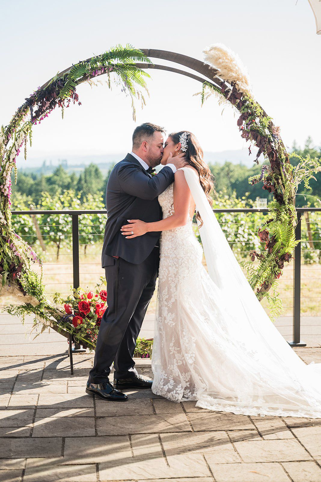 Bride and groom share first kiss under floral circular arch at outdoor vineyard wedding ceremony.