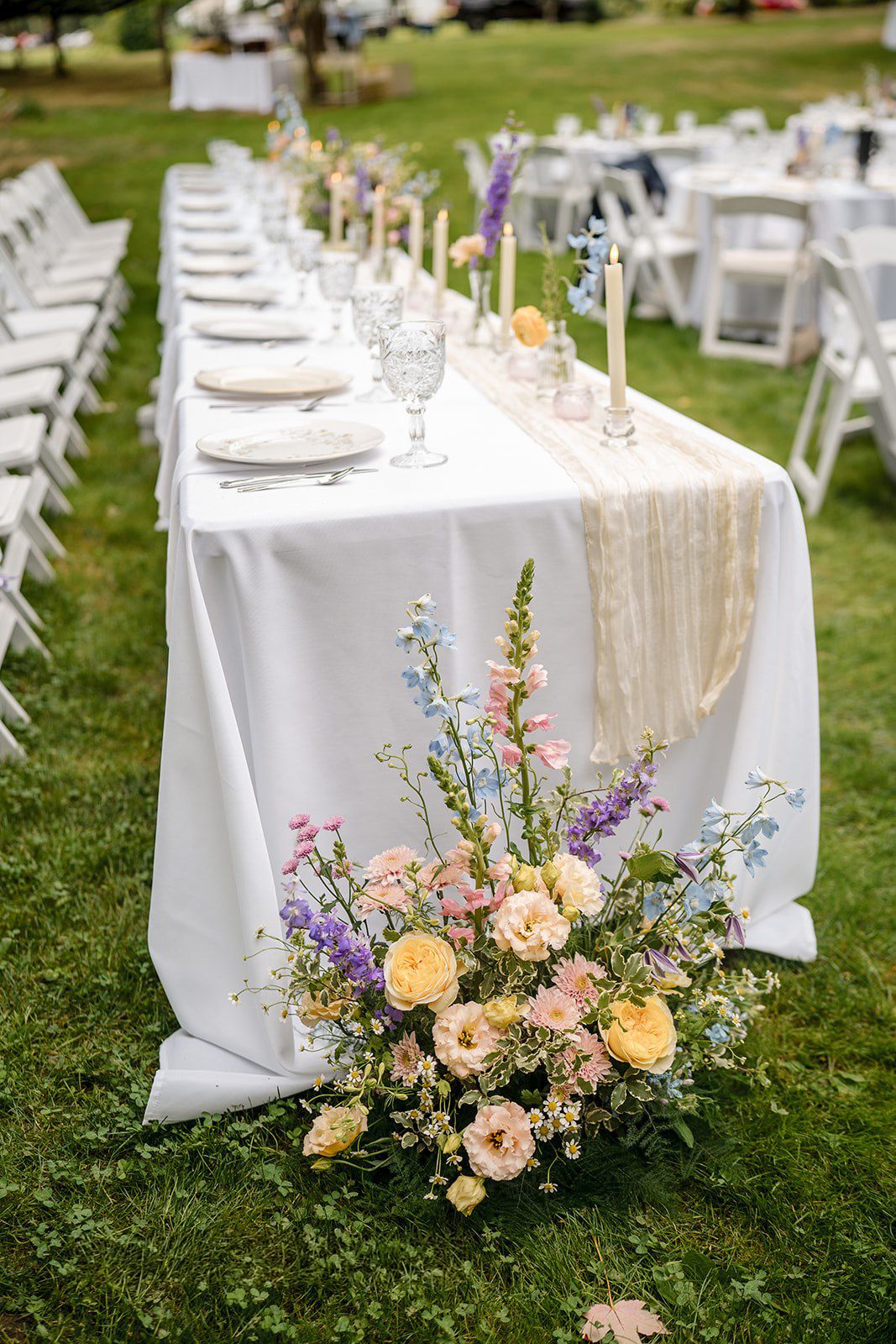 Elegant outdoor wedding reception table with white linen, pastel floral arrangement, candles, and crystal glassware on green grass.